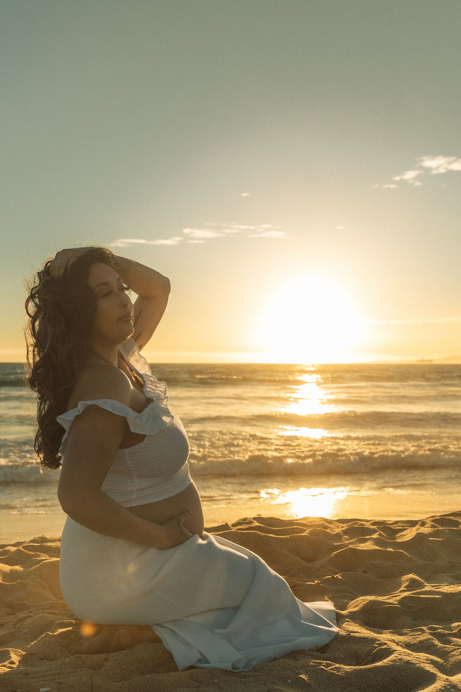 Pregnant woman in white dress sitting on sandy beach at golden sunset, cradling baby bump.