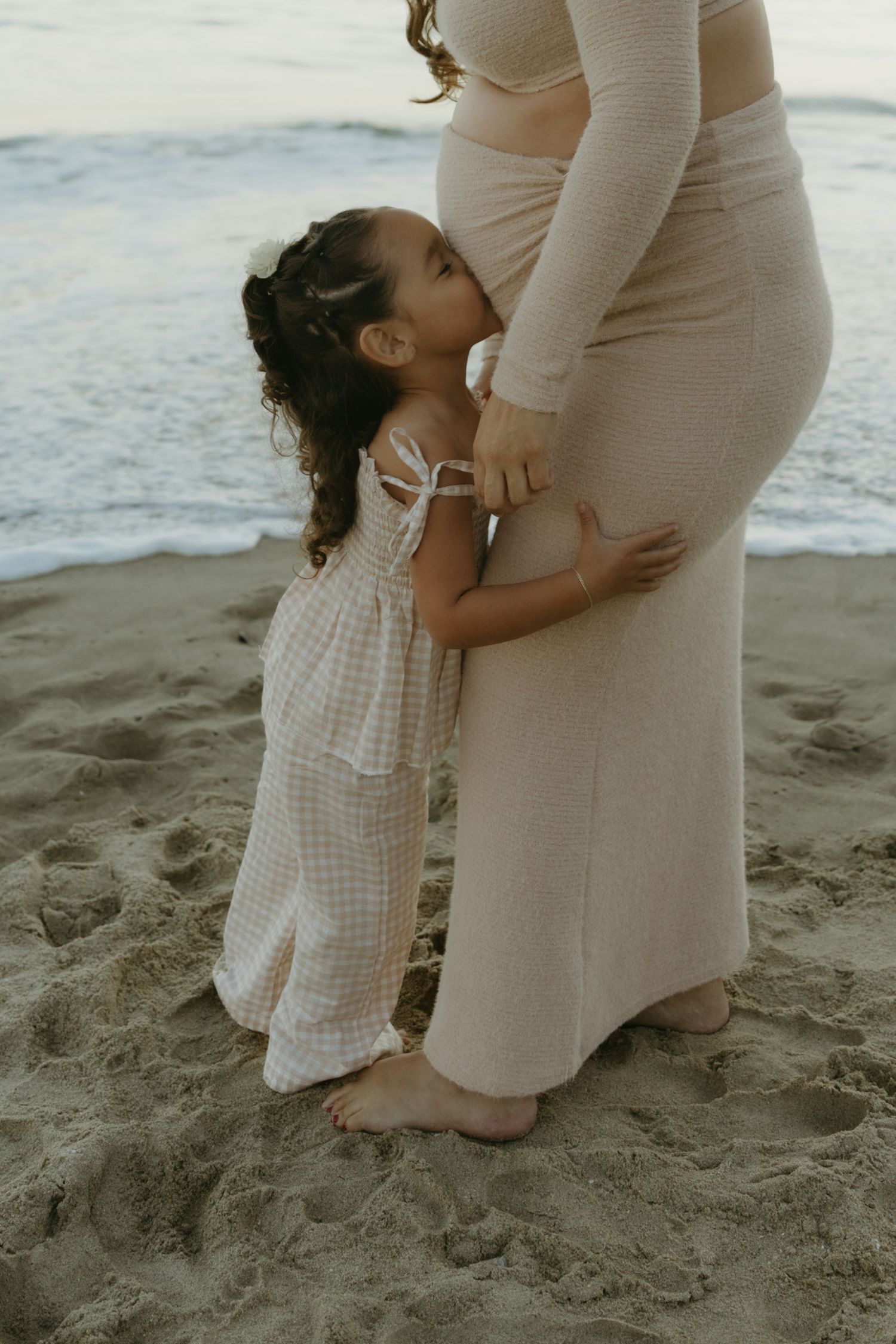 Young curly-haired girl hugs pregnant mothers belly on sandy beach near ocean waves.
