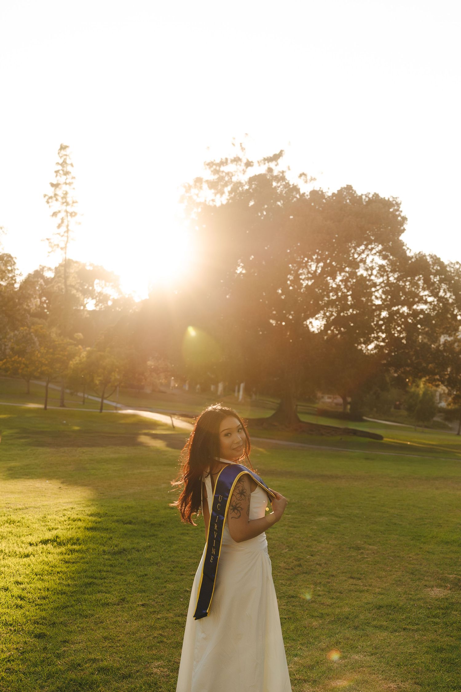 Young woman in white graduation dress and blue sash posing in sunlit park during golden hour.