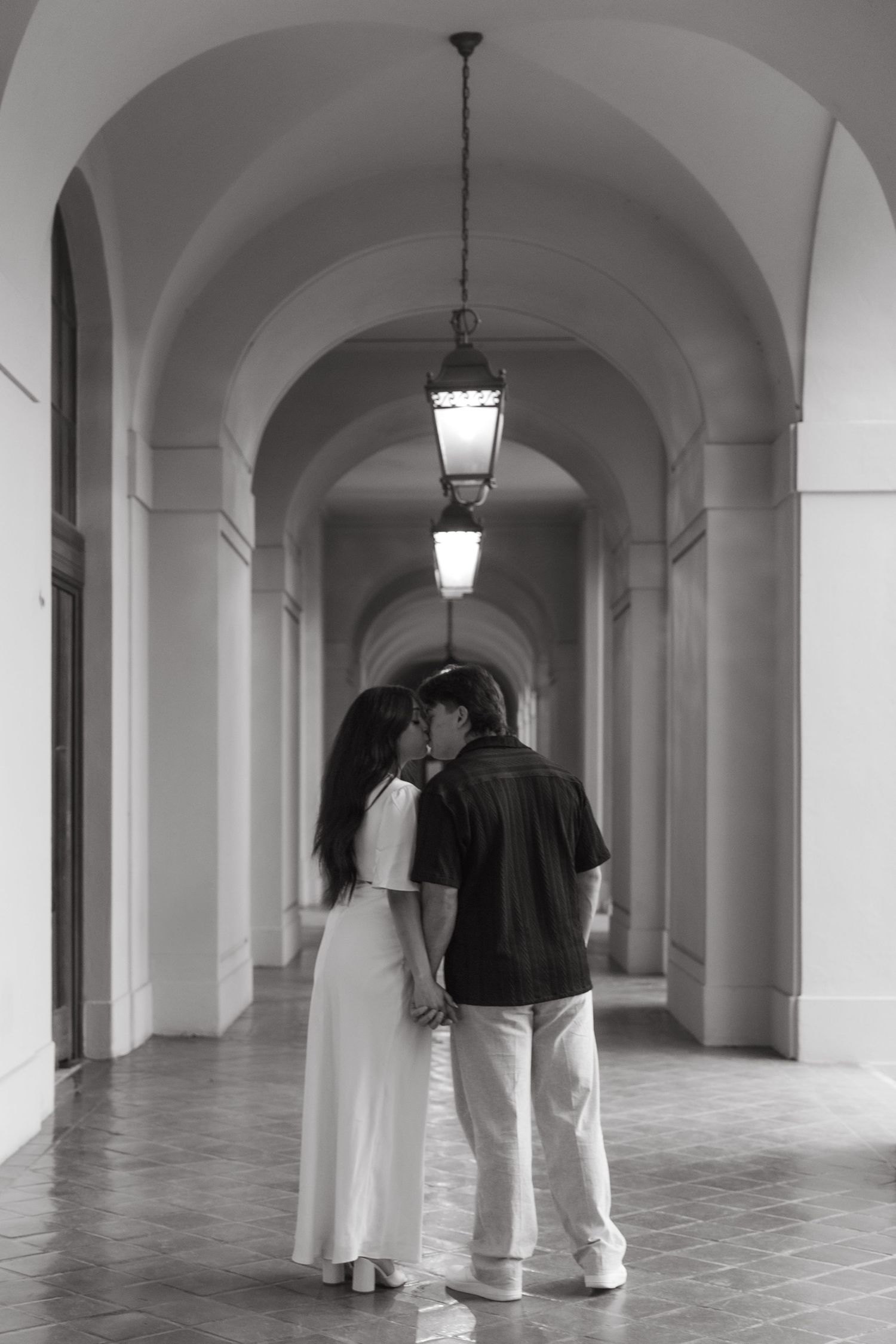 Black and white photo of couple sharing a kiss in arched corridor with hanging lanterns.