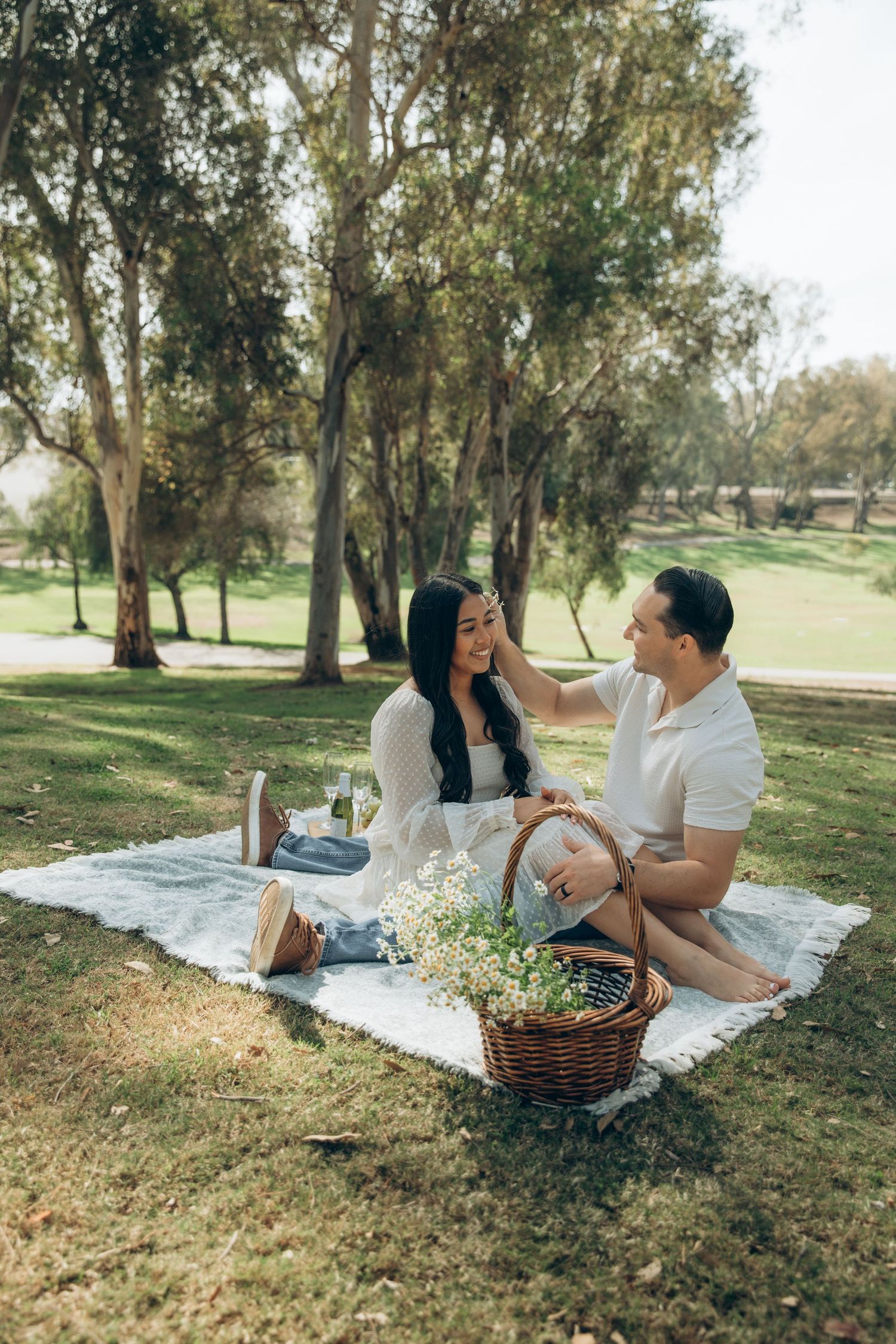 Happy couple relaxing on a picnic blanket in a park, man gently touching womans hair with flowers nearby.