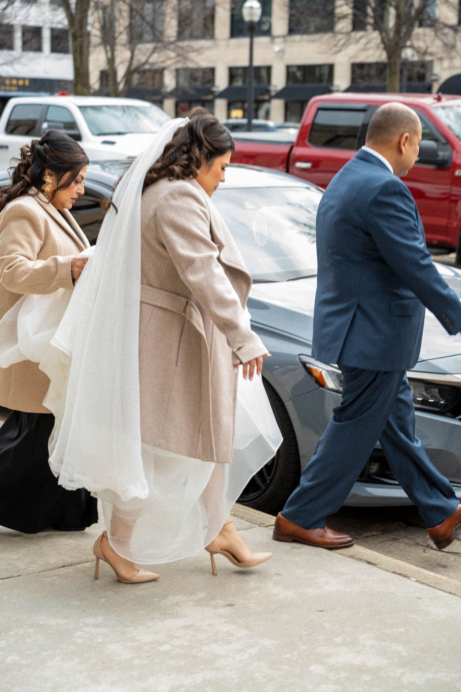 Bride in white gown and veil arriving at church with bridesmaids in beige coats on wedding day.
