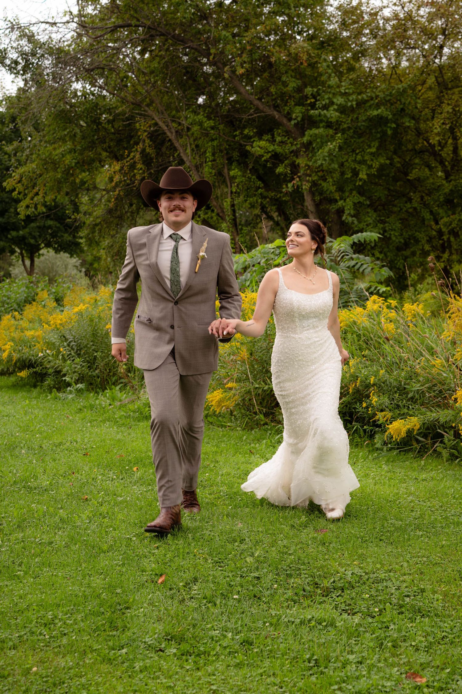 Bride and groom walk hand-in-hand through lush garden, groom in tan suit and cowboy hat, bride in fitted white gown.
