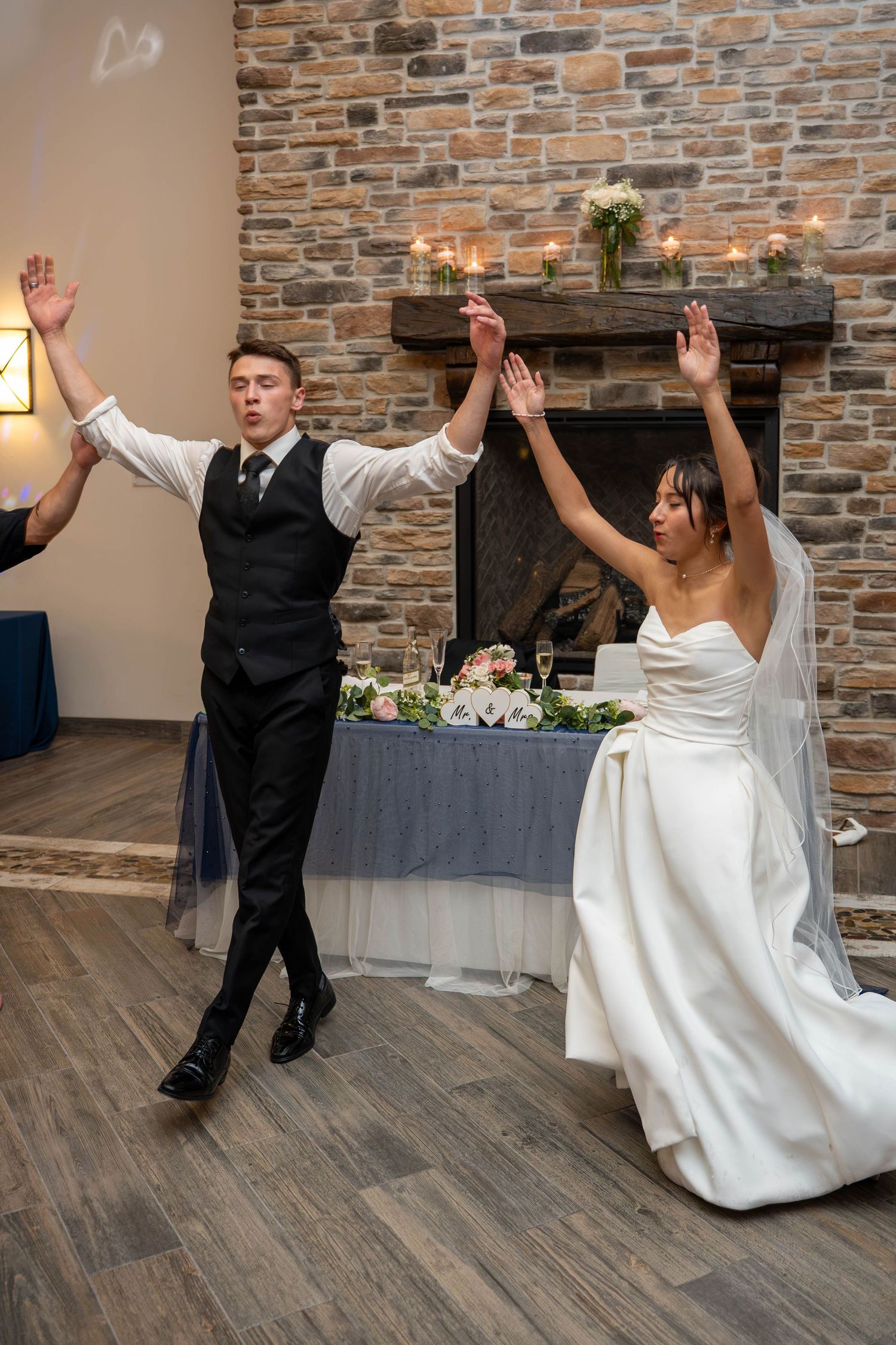Bride and groom joyfully raise their arms during wedding reception dance near stone fireplace.