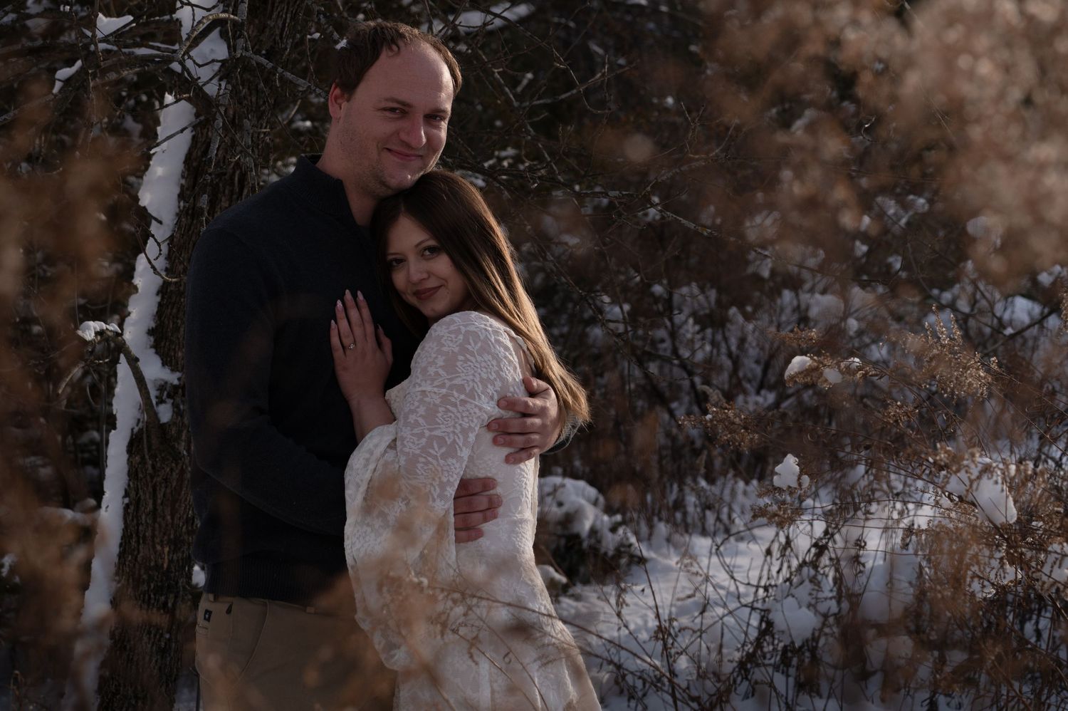 Couple embracing in snowy winter forest, woman in white lace dress and man in black sweater.