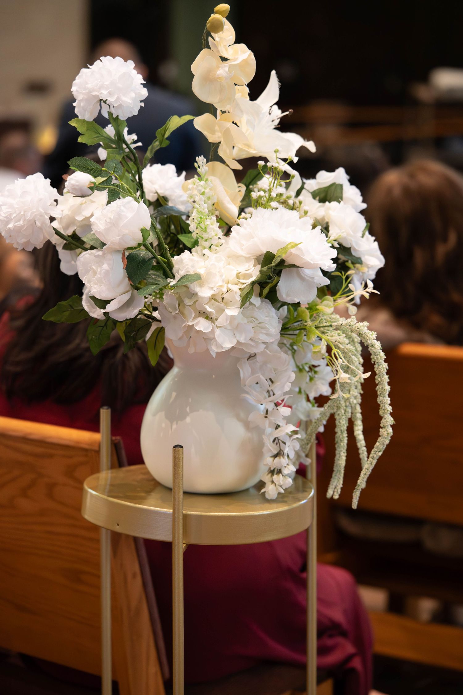 White floral arrangement in ceramic vase on gold stand decorating church aisle for wedding.