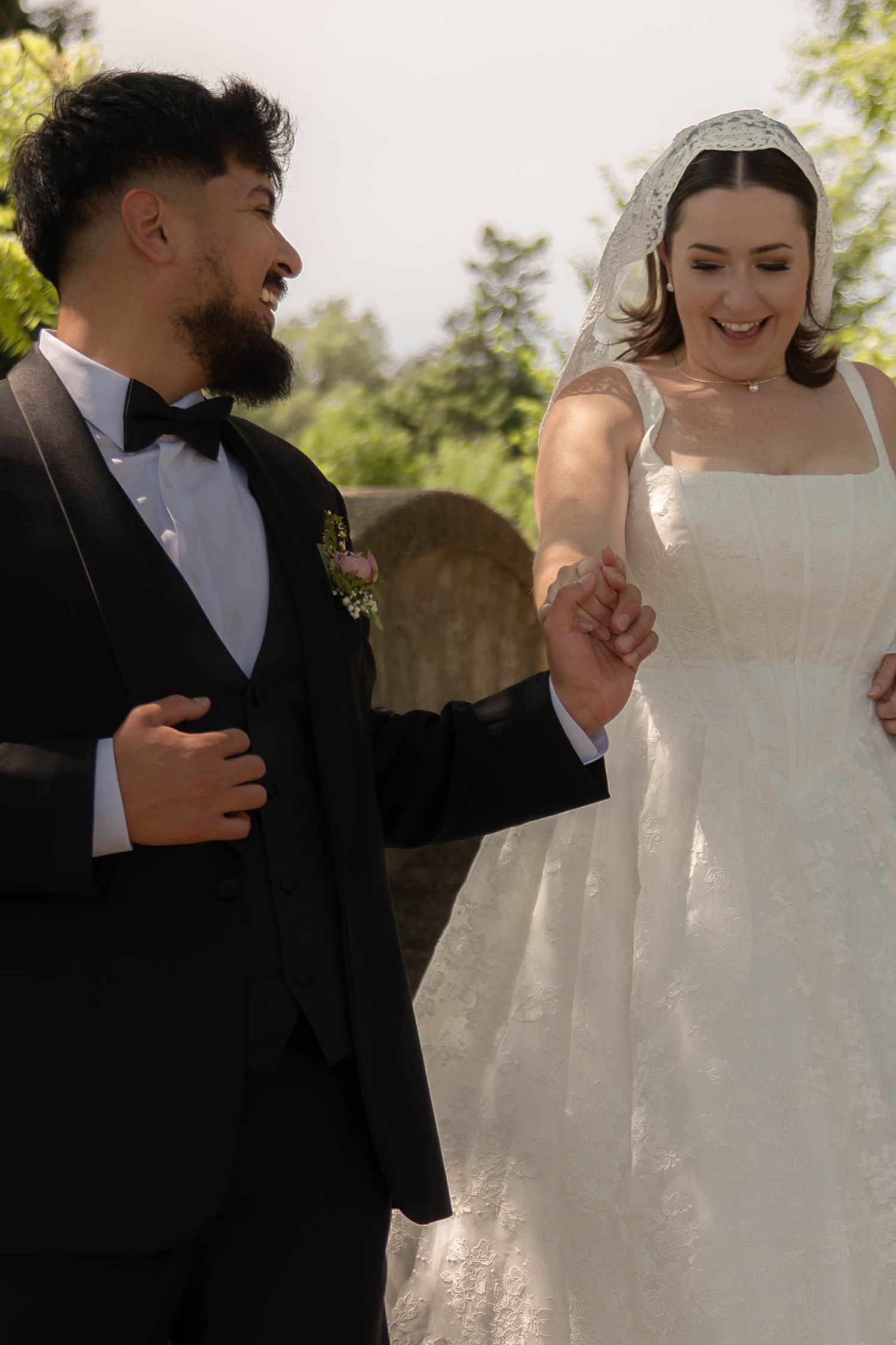 Smiling bride in lace veil and groom in black bow tie tuxedo walk hand in hand outdoors after wedding.
