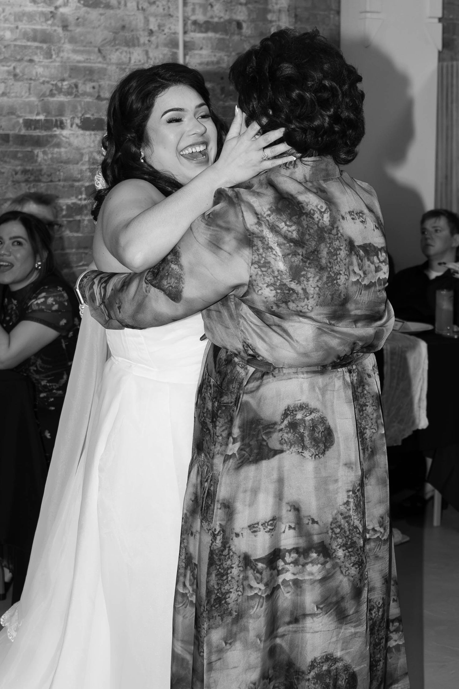 Joyful bride in white gown hugging a guest during wedding reception in black and white photo.