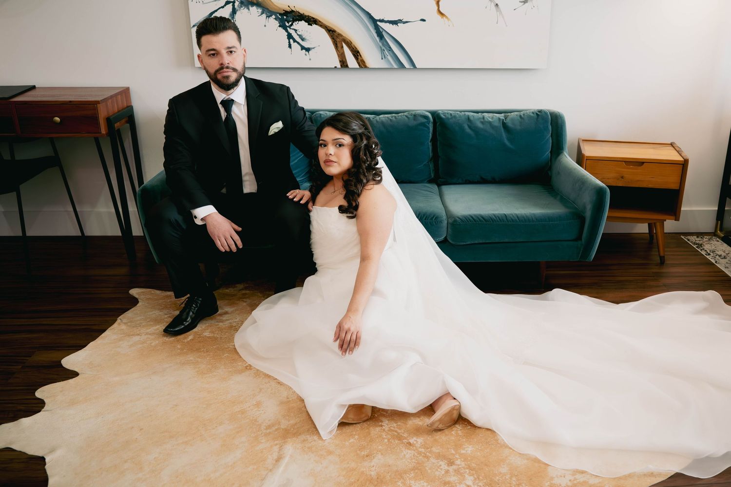 Bride and groom pose elegantly in a modern room, bride seated on floor with flowing white gown spread out.