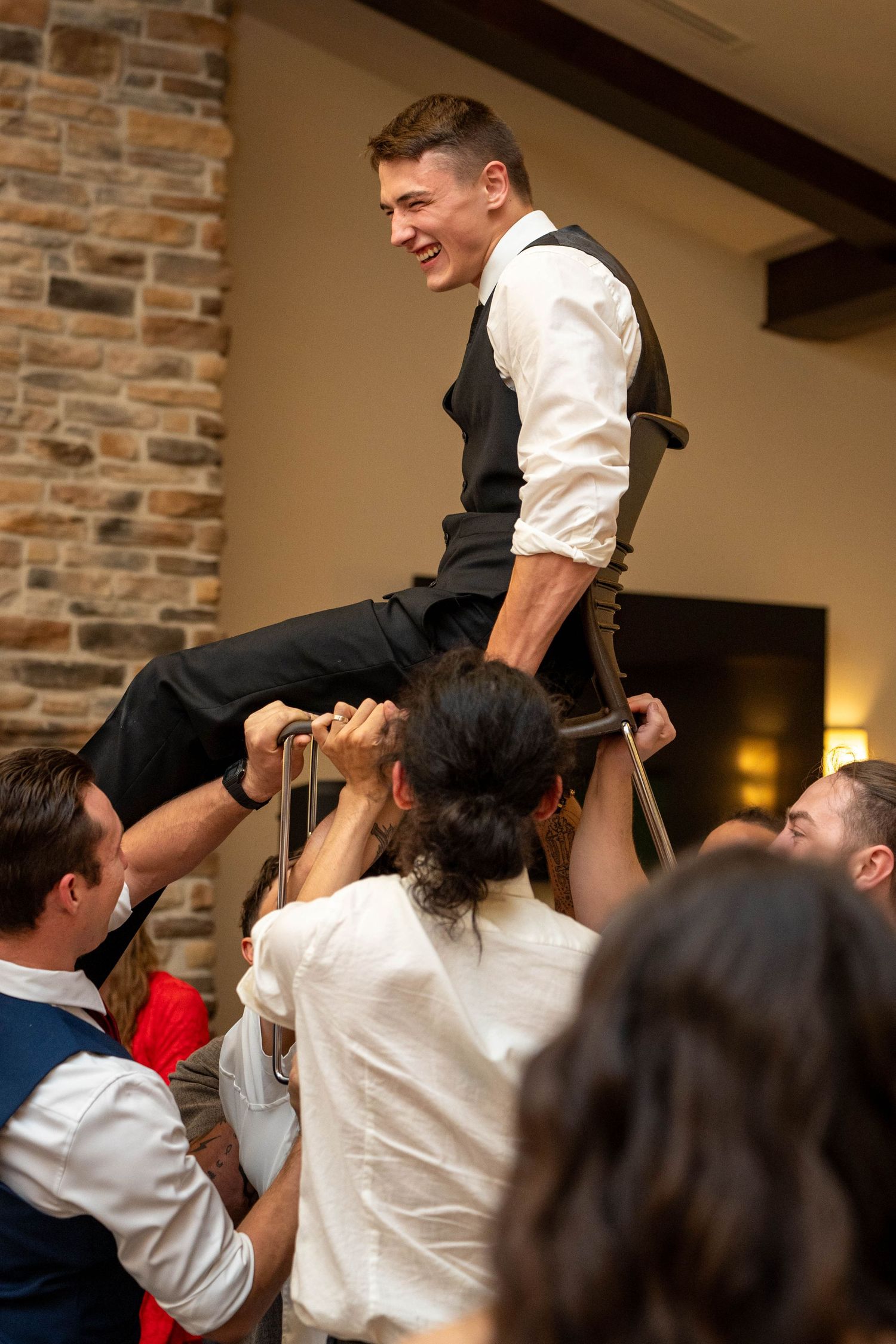 Groom laughing while being lifted on chair by groomsmen during wedding reception celebration.