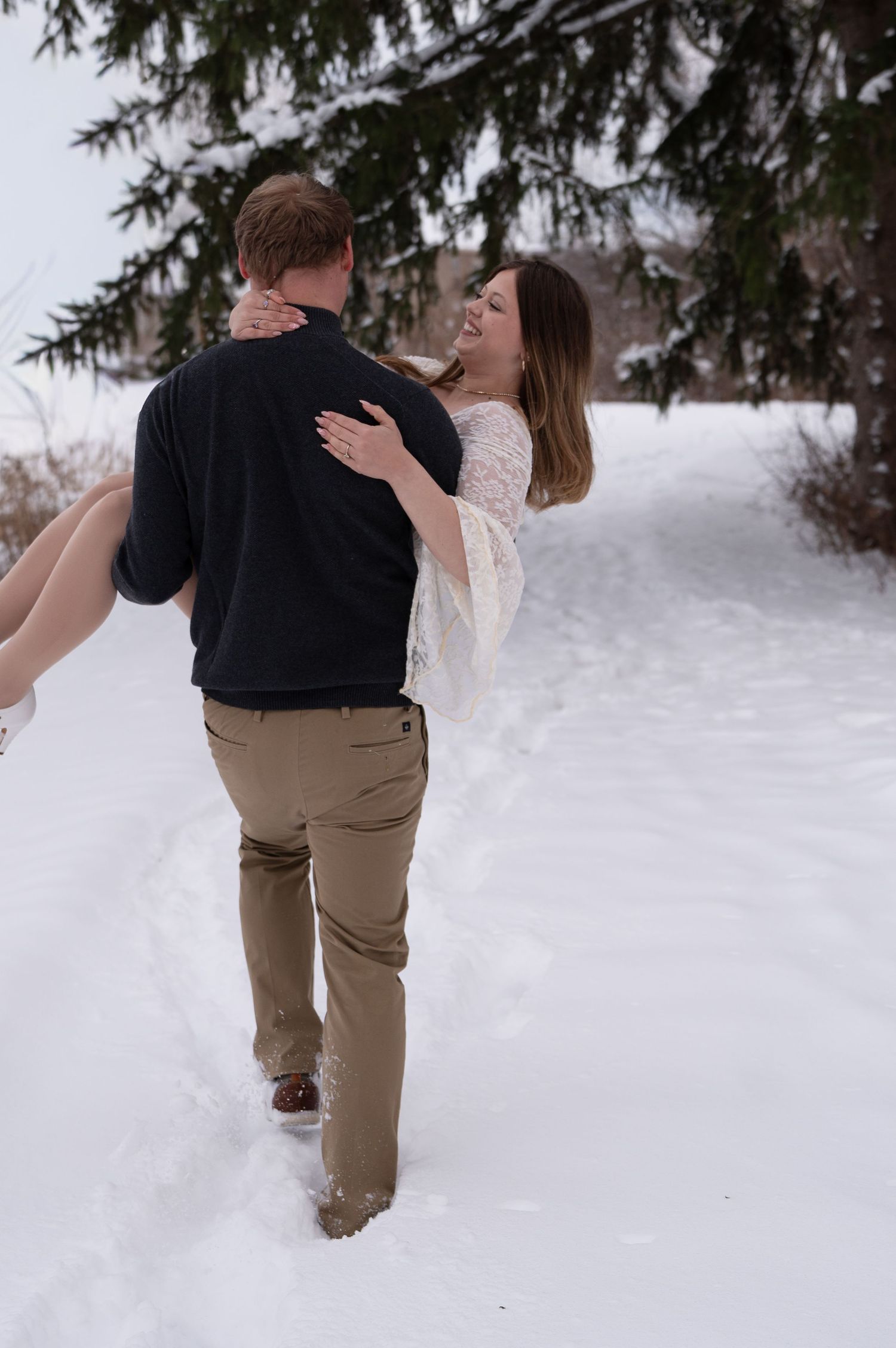 Groom carries laughing bride through a snowy forest path lined with evergreen trees.