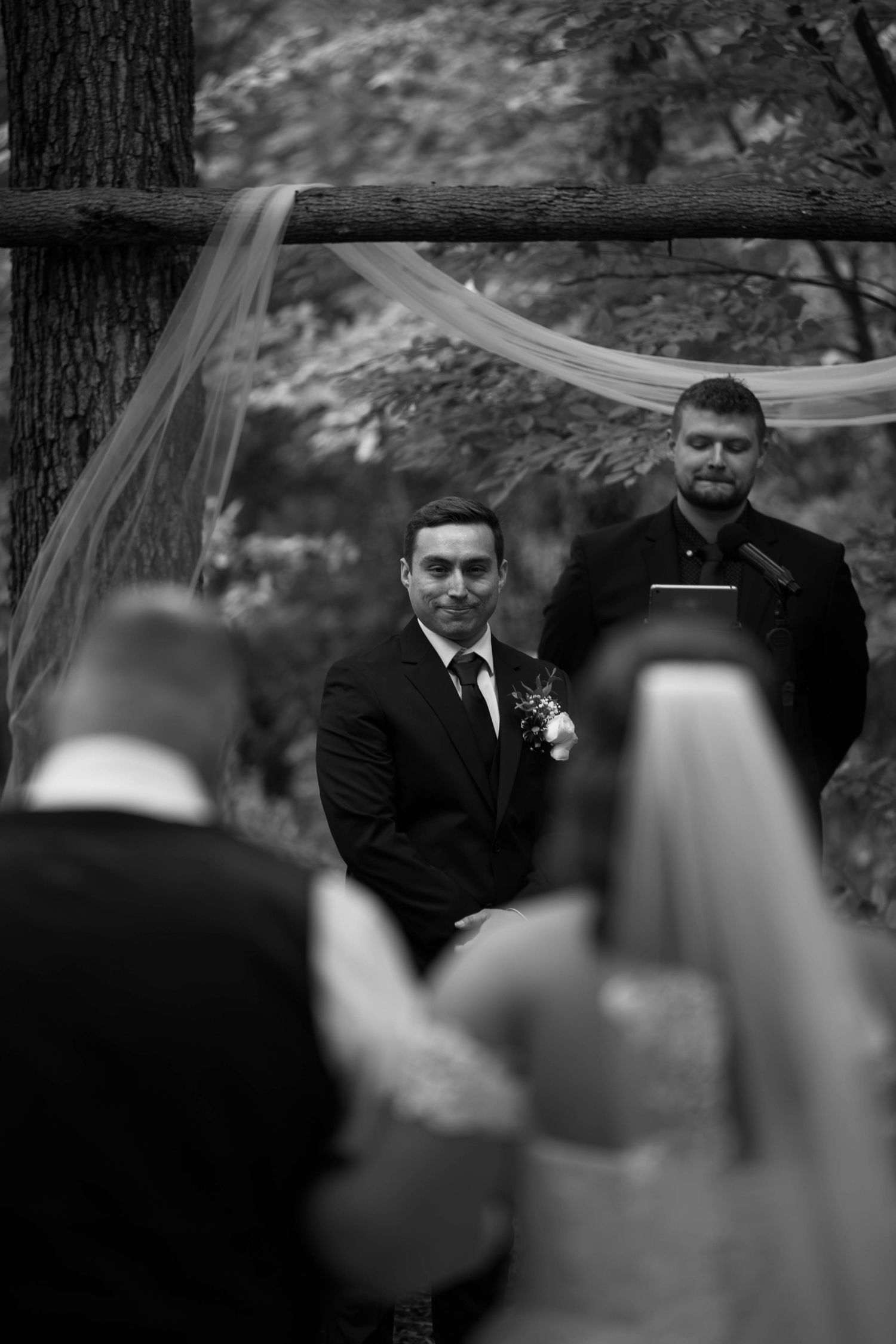 Black and white wedding ceremony photo showing groom awaiting bride at rustic woodland altar with officiant standing behind.