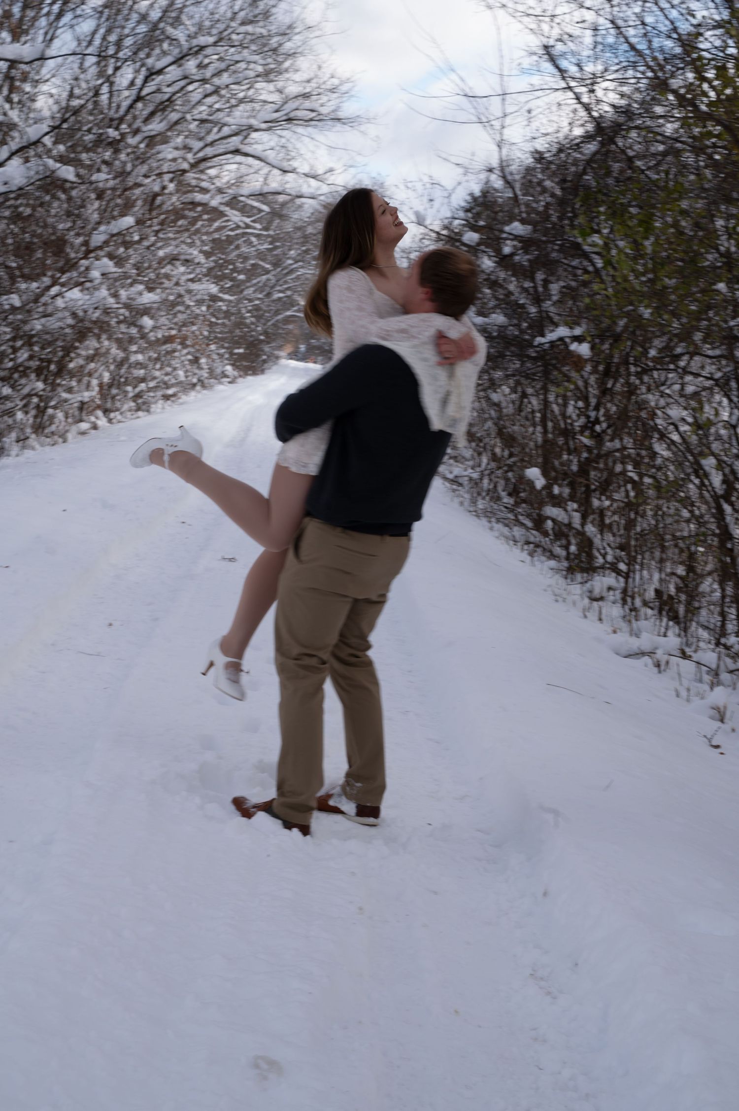 Groom lifting bride in white dress on snow-covered path surrounded by winter trees.