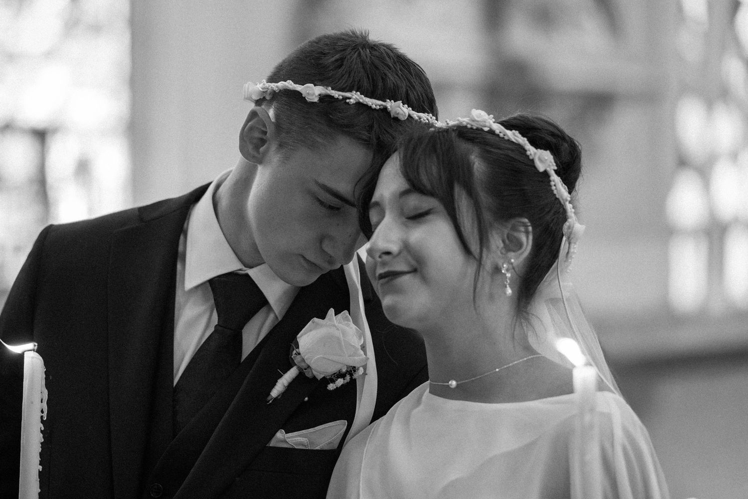 Tender black and white portrait of bride and groom wearing floral crowns during church ceremony.