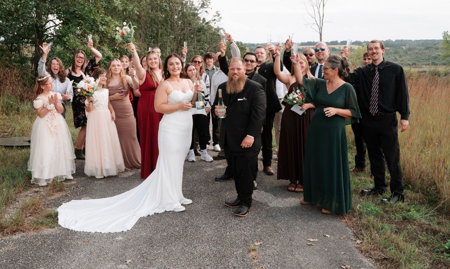 Bride and groom celebrating with large wedding party on scenic outdoor pathway.