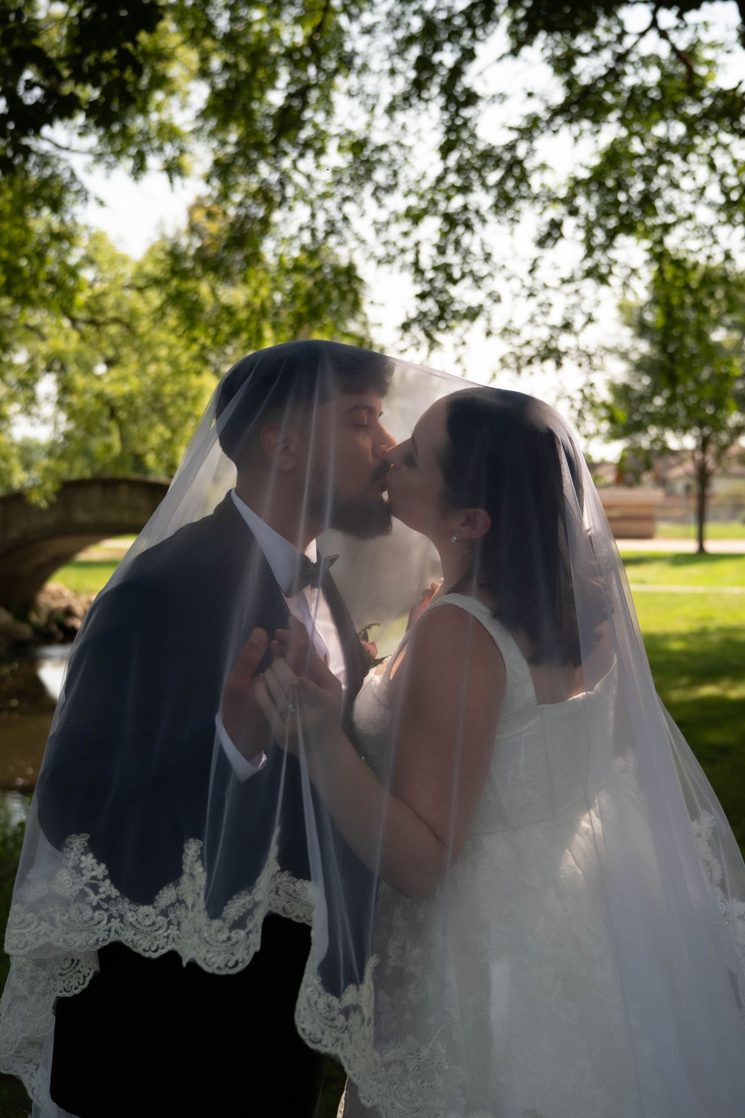 Bride and groom share a kiss beneath flowing lace wedding veil in a sunlit park with green trees.