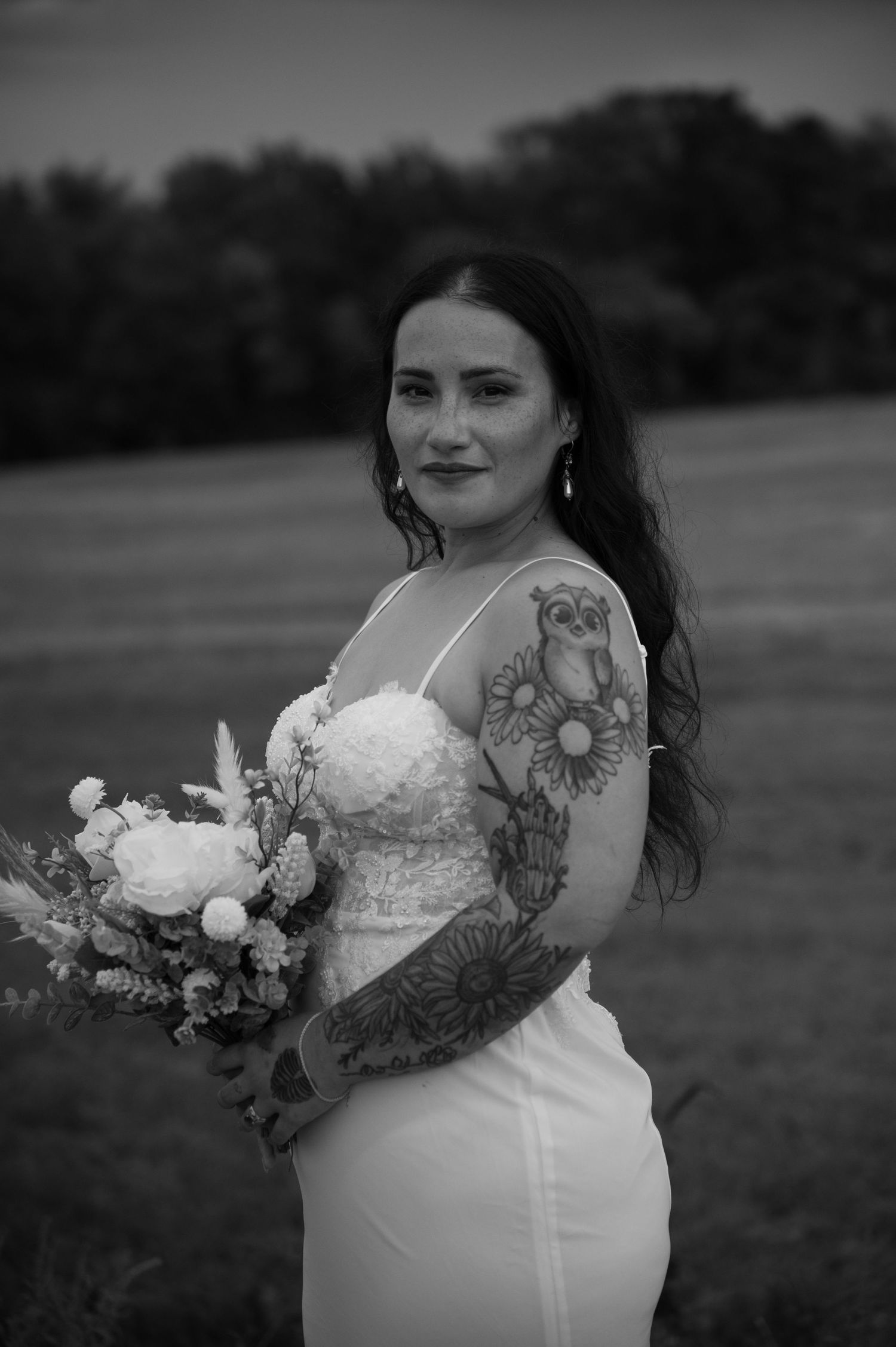 Black and white photo of a tattooed bride holding a floral bouquet in a white gown outdoors.
