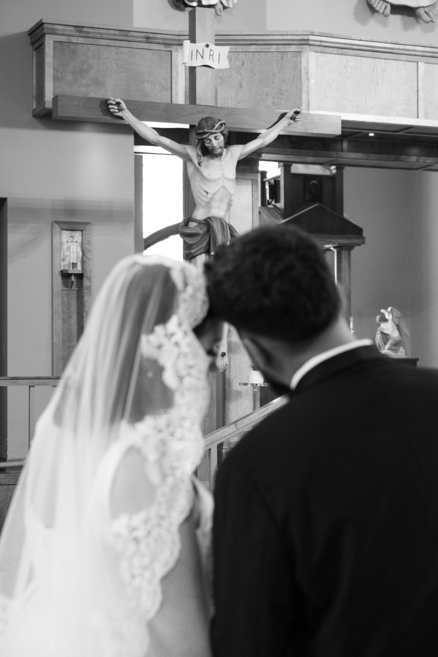 Black and white photo of bride and groom bowing before a wooden crucifix during a Catholic wedding ceremony.