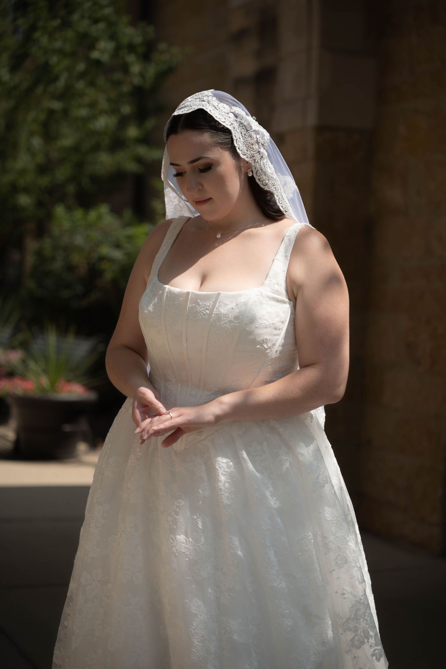 Elegant bride in sleeveless white lace gown and lace veil stands outdoors looking down with clasped hands.