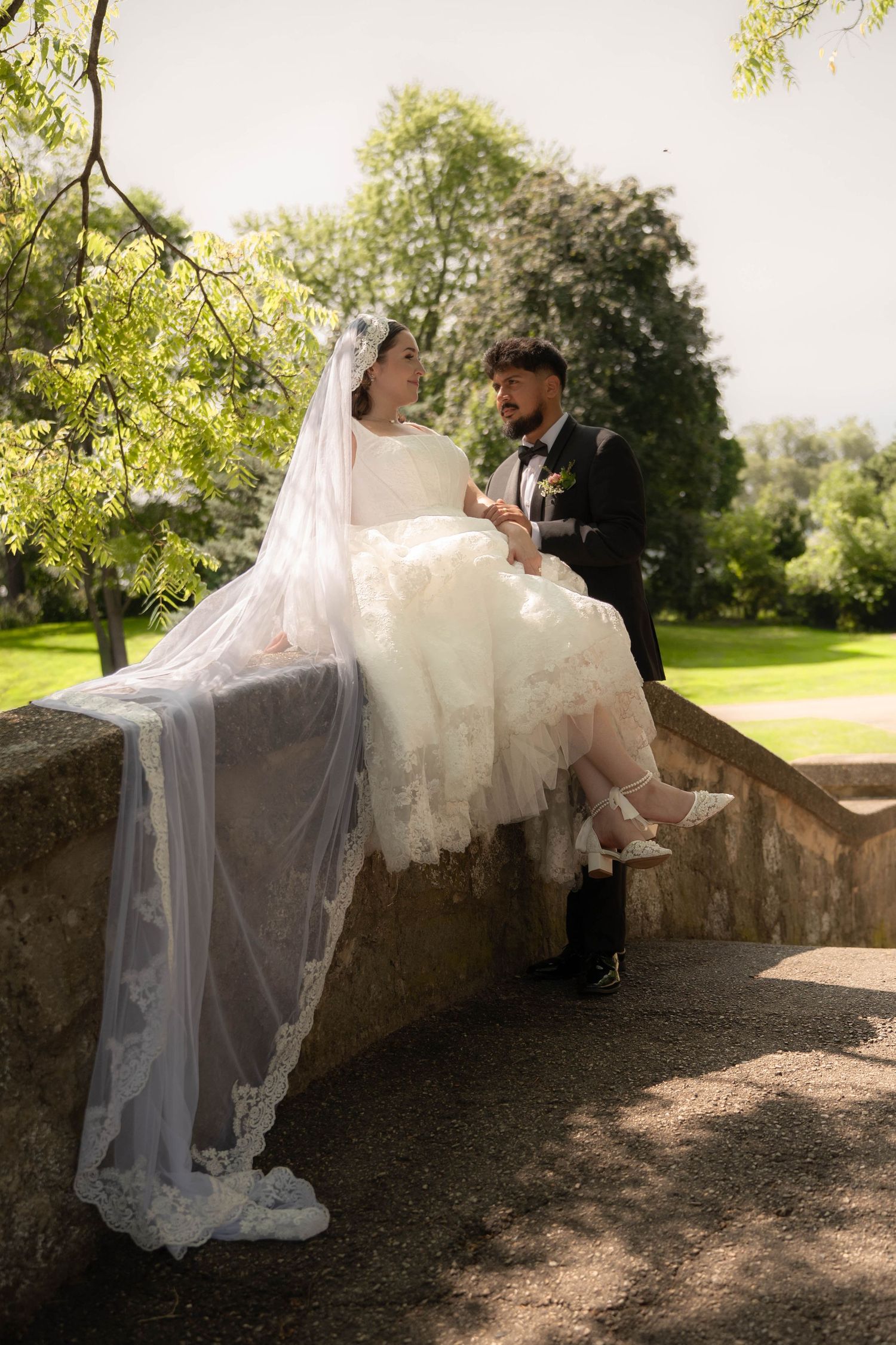 Bride in lace wedding gown and veil sits on stone wall while groom in black tux holds her hands outdoors.