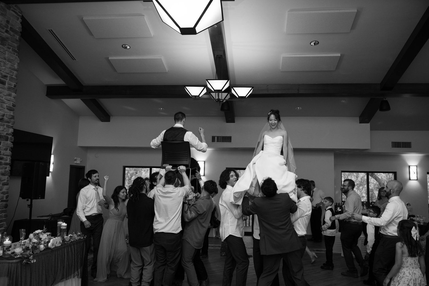 Black and white photo of bride and groom being lifted by guests at reception.