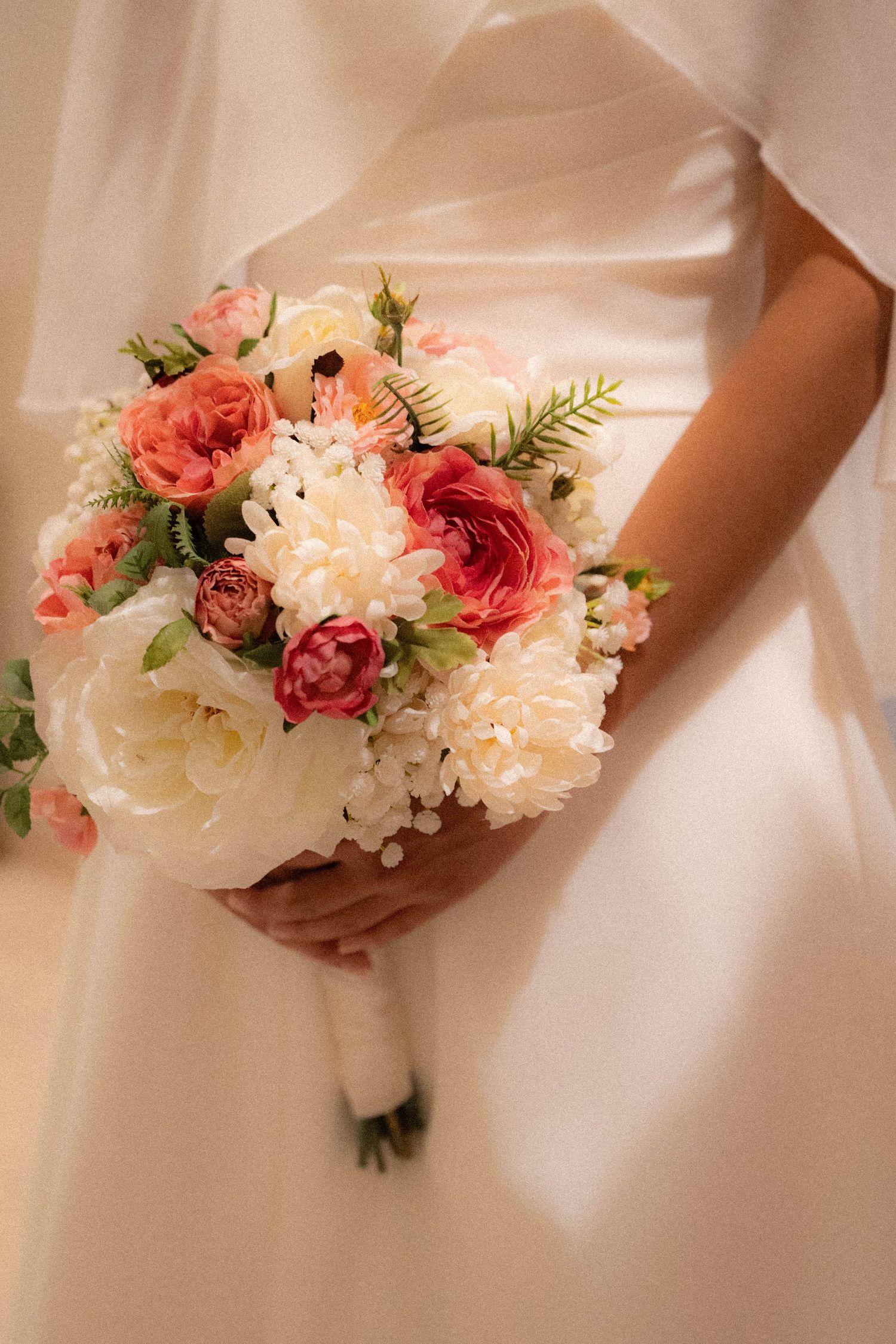 Bride in white satin gown holds elegant bouquet of pink roses, peonies, and white chrysanthemums.