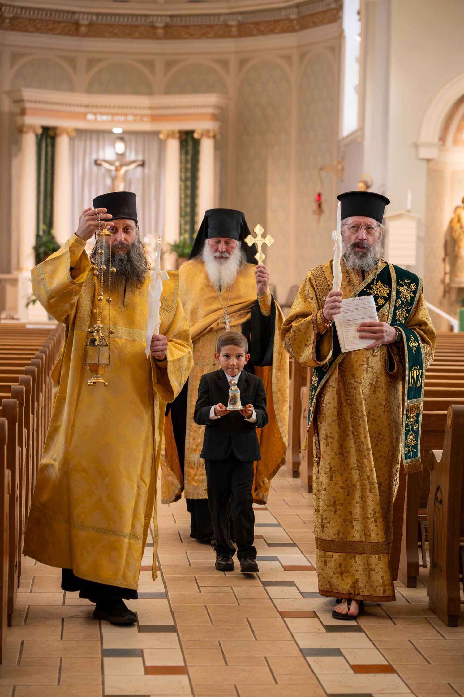 Three Orthodox priests in golden vestments processing down church aisle with young boy during wedding.