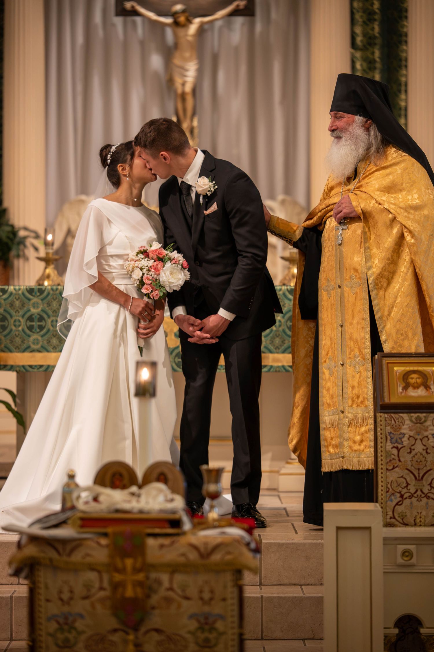 Bride and groom share first kiss at altar during Orthodox Christian wedding ceremony with priest.