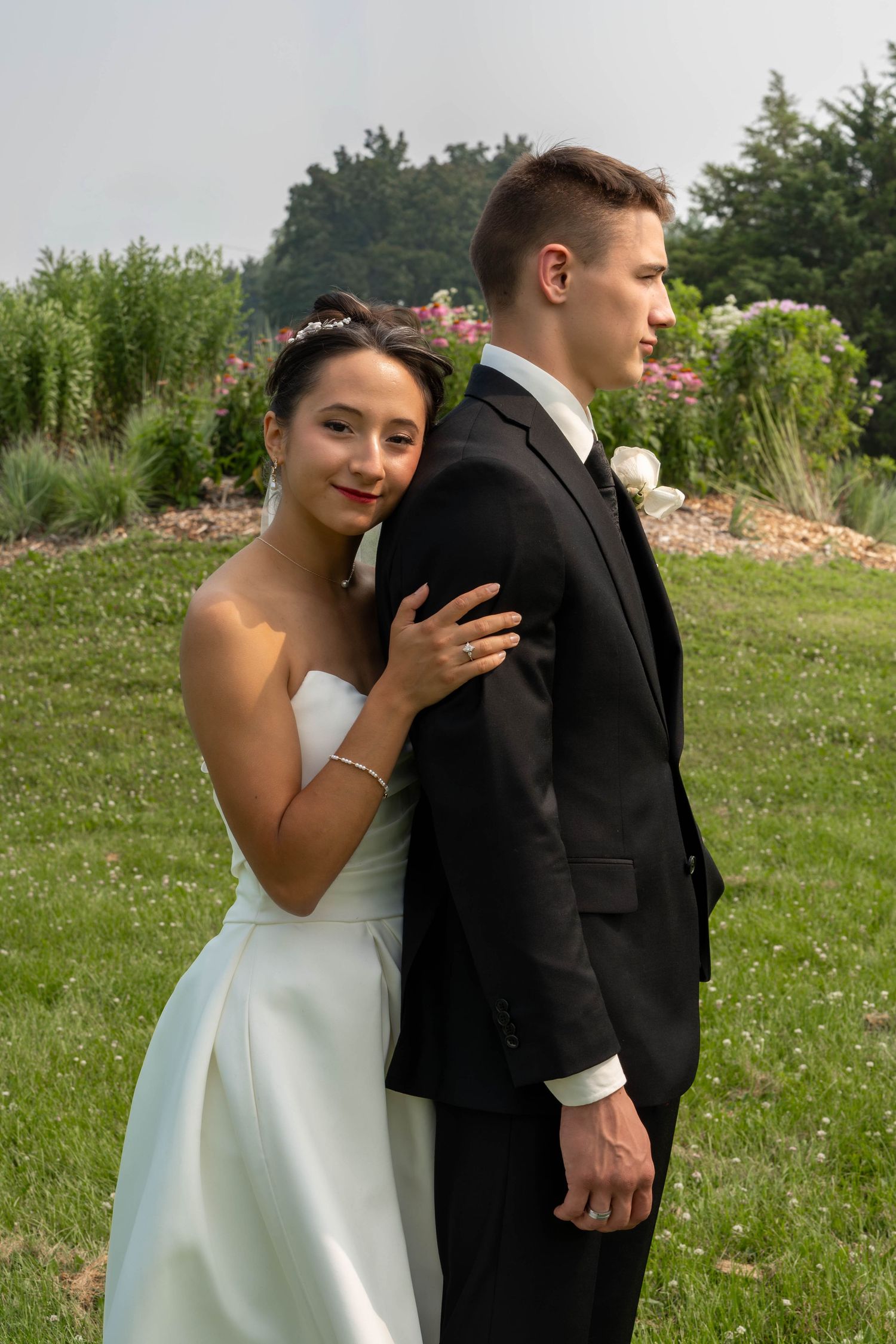 Bride in white strapless gown leaning on grooms shoulder outdoors in lush green garden setting.
