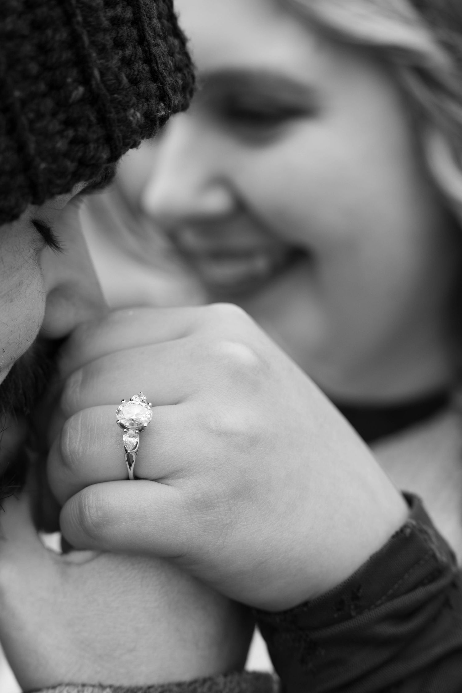 Black and white close-up of engagement ring on womans hand while couple embraces.