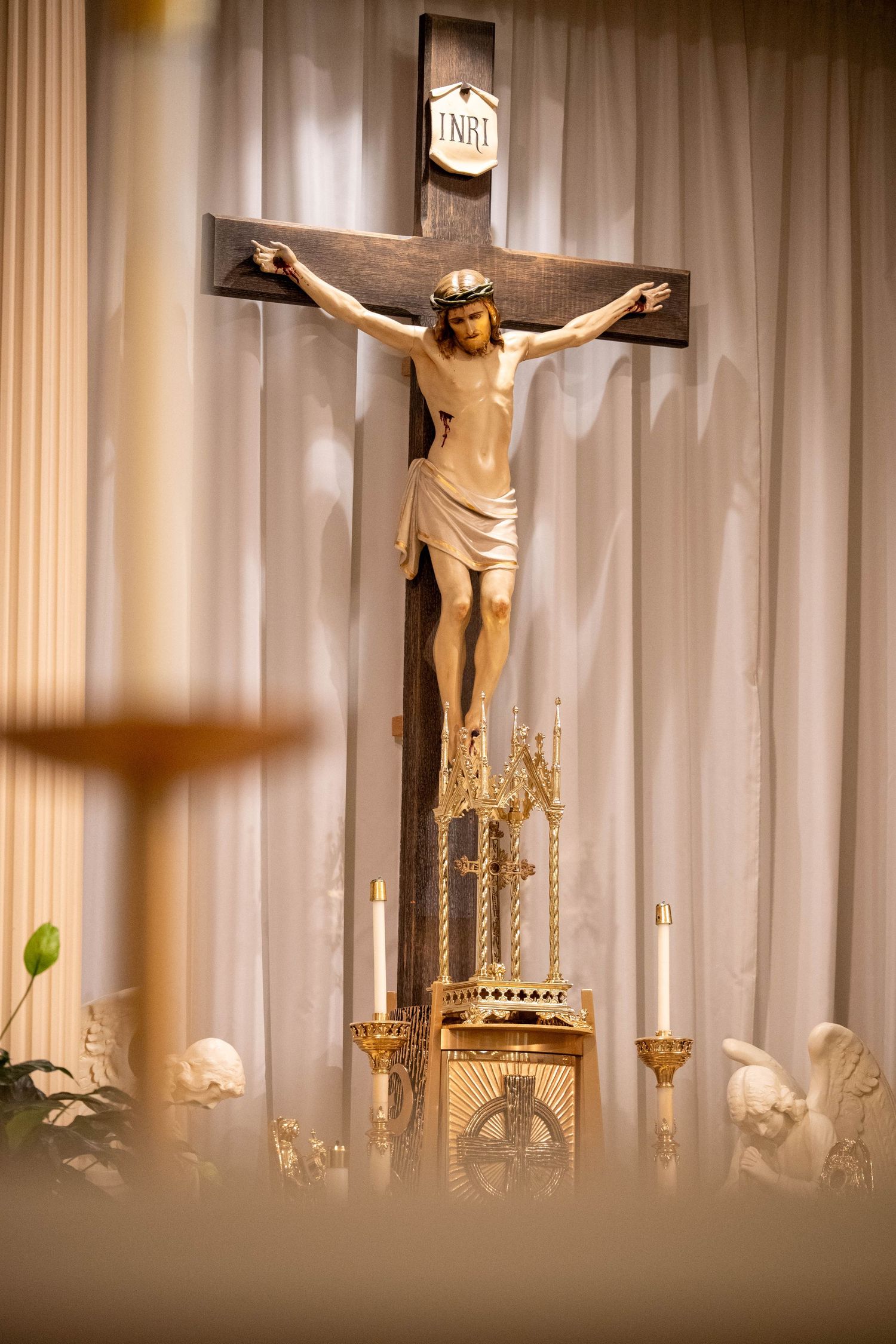 A crucifix with INRI inscription displayed on a church altar with golden candleholders and white curtains.