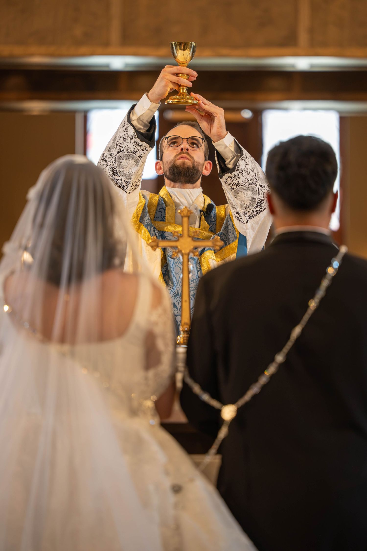 Orthodox priest raises golden chalice during wedding ceremony with bride and groom at altar.