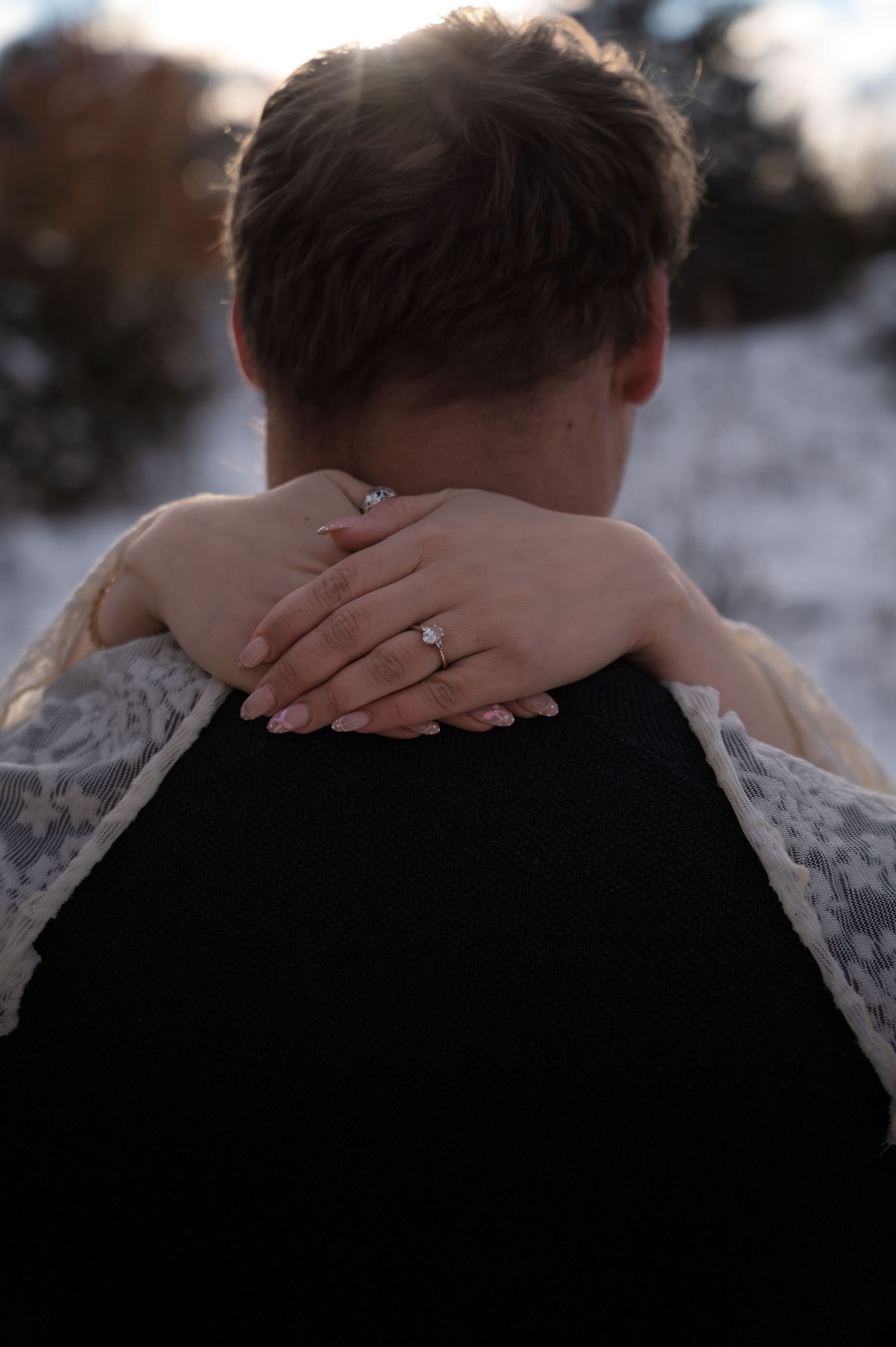 Bride rests her hand on groom's shoulder, showcasing her diamond engagement ring in snow.