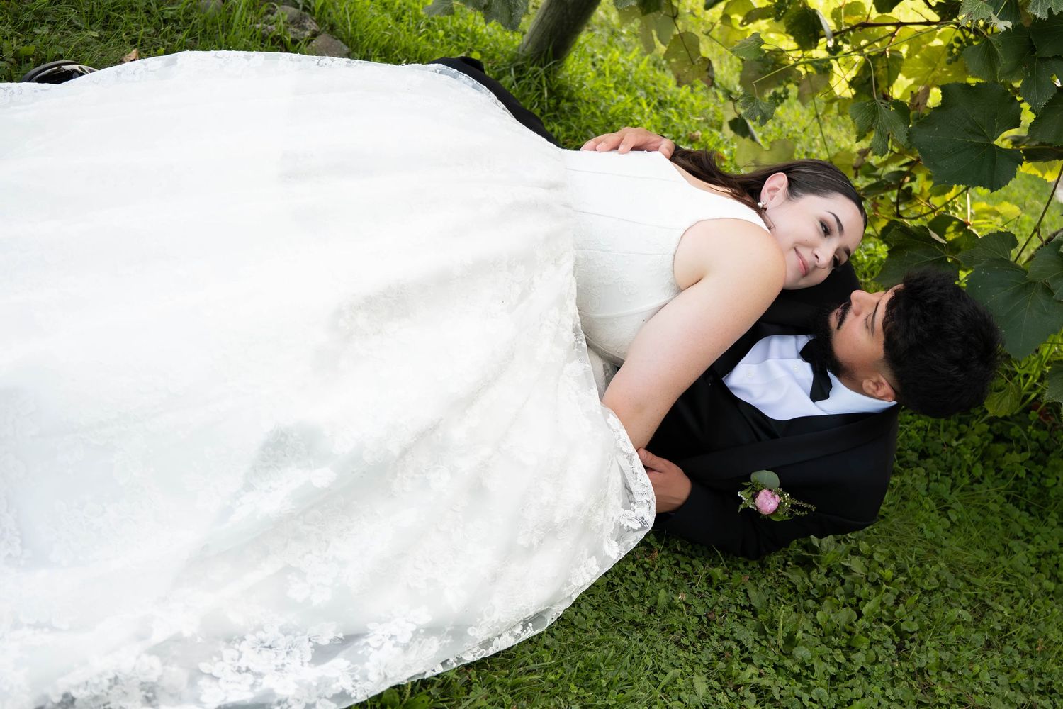 Bride and groom lie together on lush green grass, sharing a romantic embrace in wedding attire.