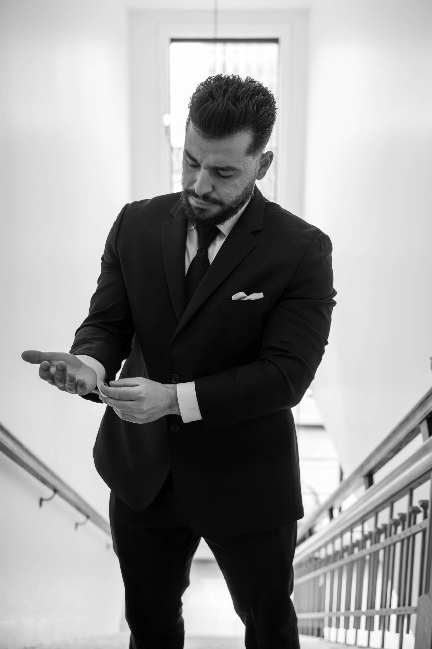 Dapper groom in black suit checks phone on staircase in elegant black and white photo.