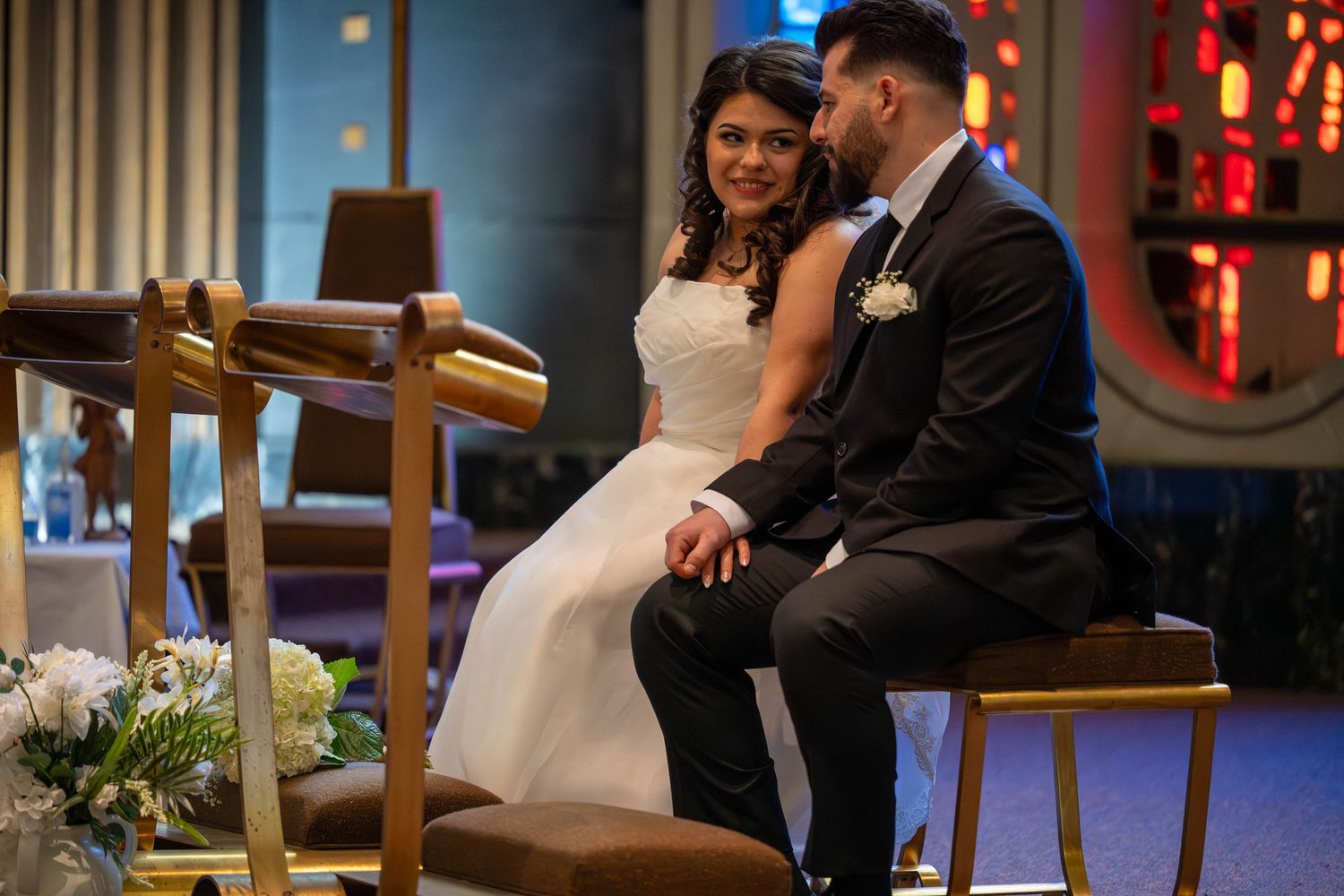 Bride and groom sitting together at wedding ceremony altar, smiling lovingly at each other in modern church.