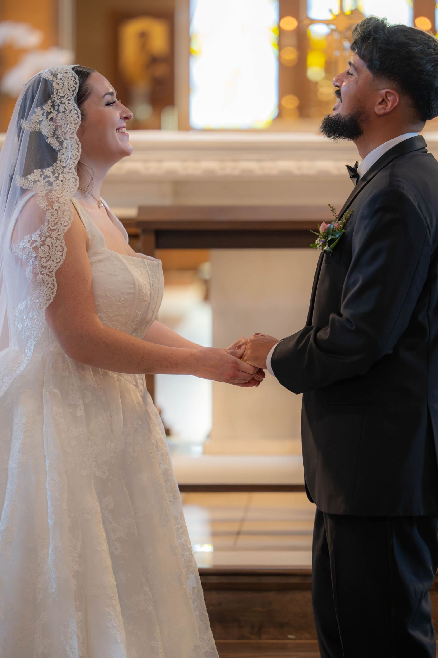 Bride in lace veil and groom in tuxedo hold hands and smile joyfully during their church wedding ceremony.
