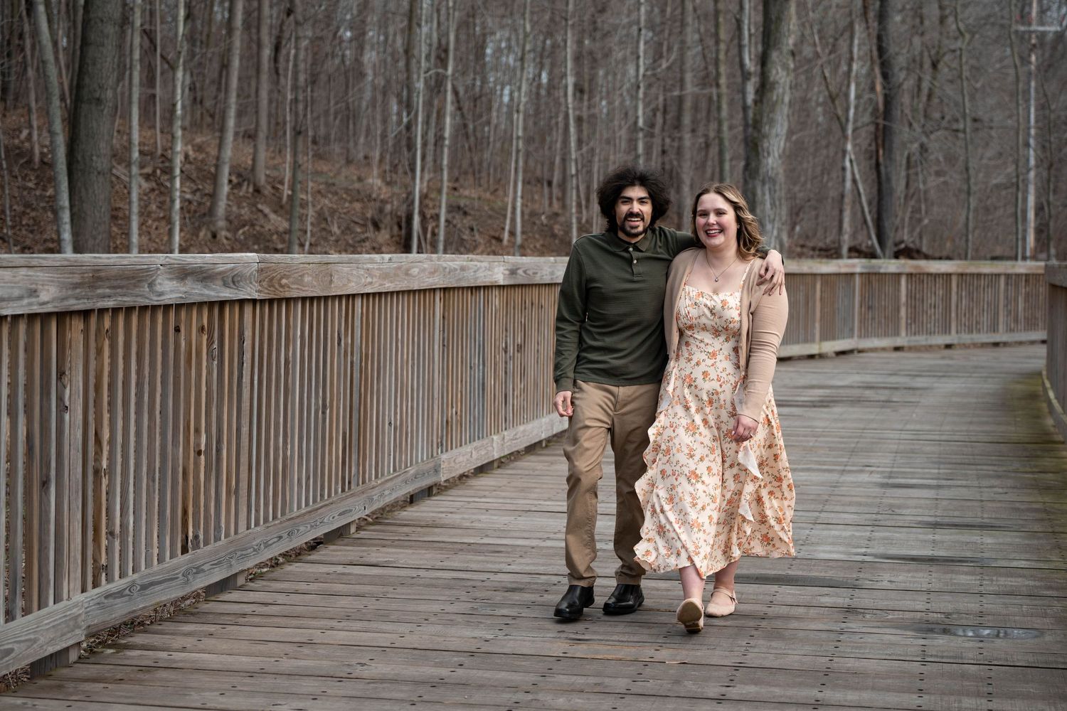 Happy couple walking hand in hand on wooden bridge surrounded by bare winter trees.