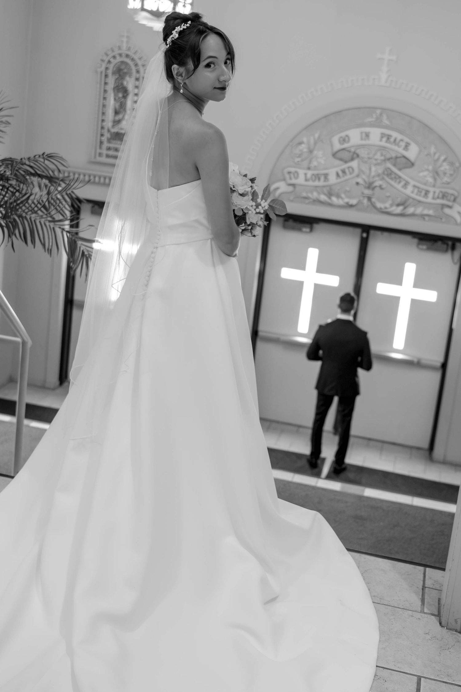 Black and white photo of bride in strapless gown with veil looking back toward church doors.