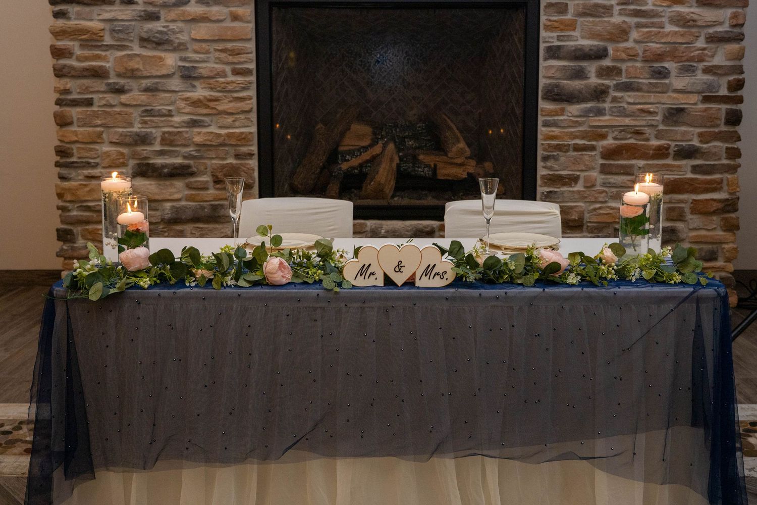 Elegant wedding sweetheart table with candles, greenery garland, and Mr. Mrs. signs by fireplace.