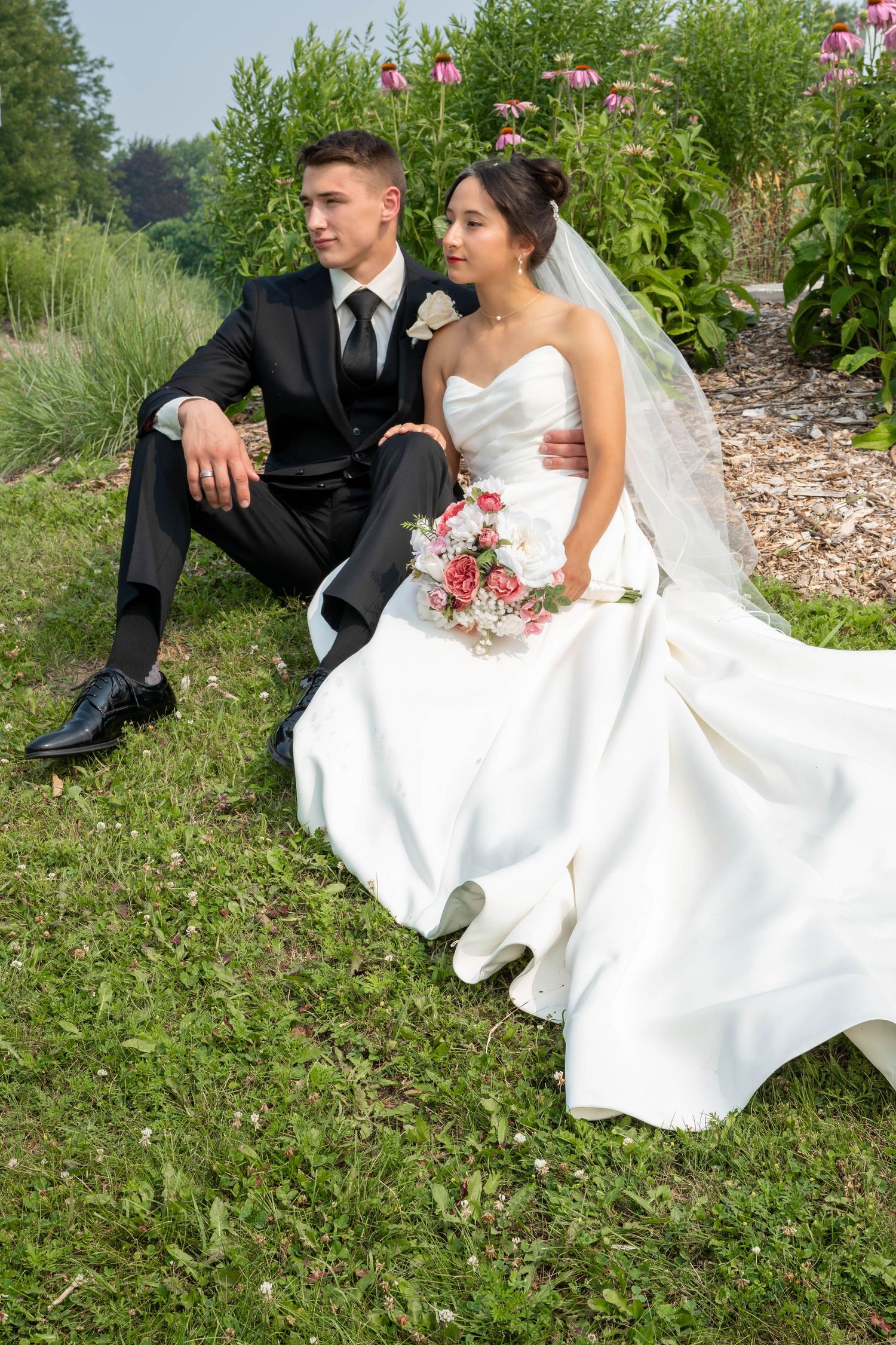 Bride in white gown holding bouquet sits with groom in black suit on grass in garden.