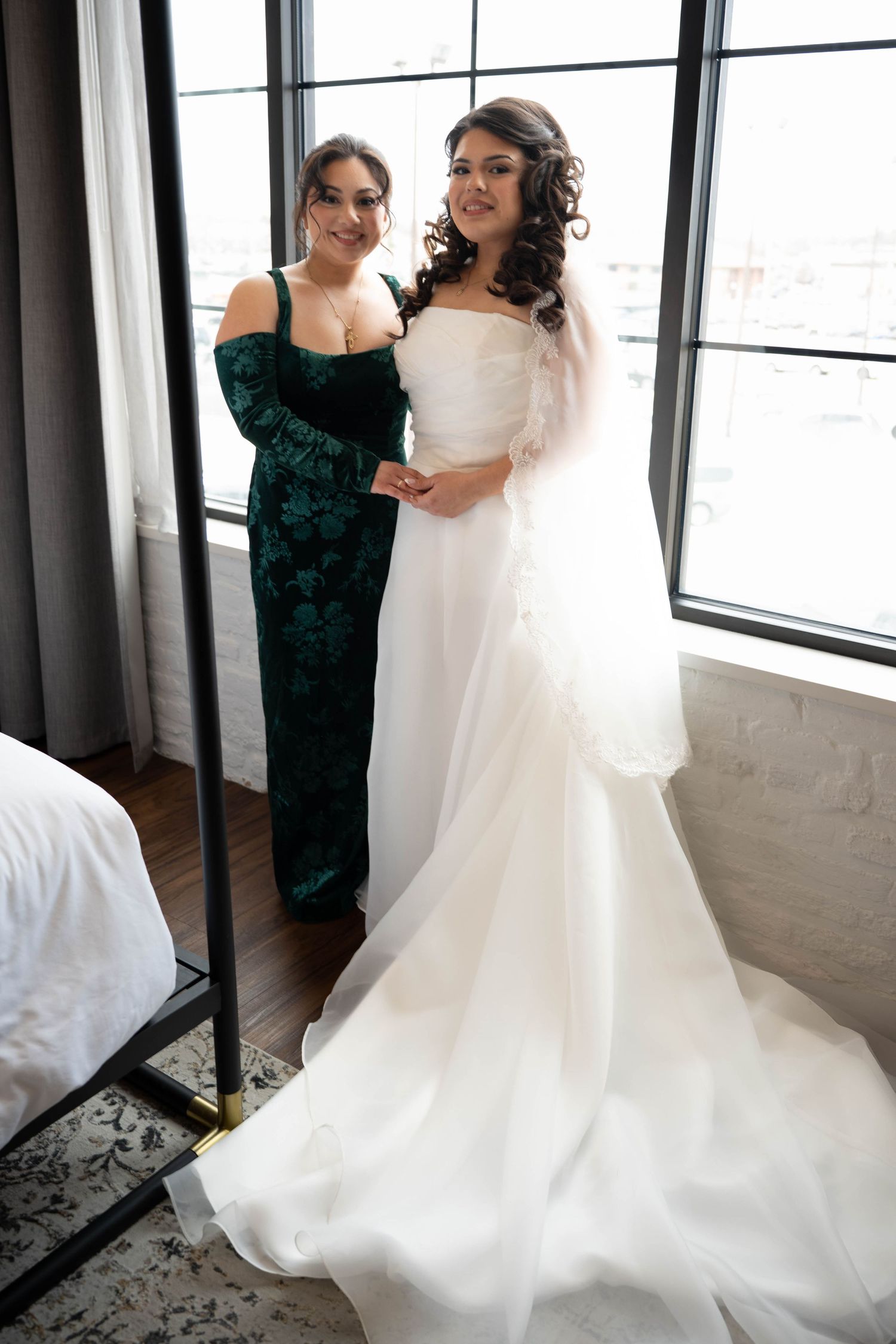 Bridesmaid in green velvet dress helps bride in white gown prepare near large windows.