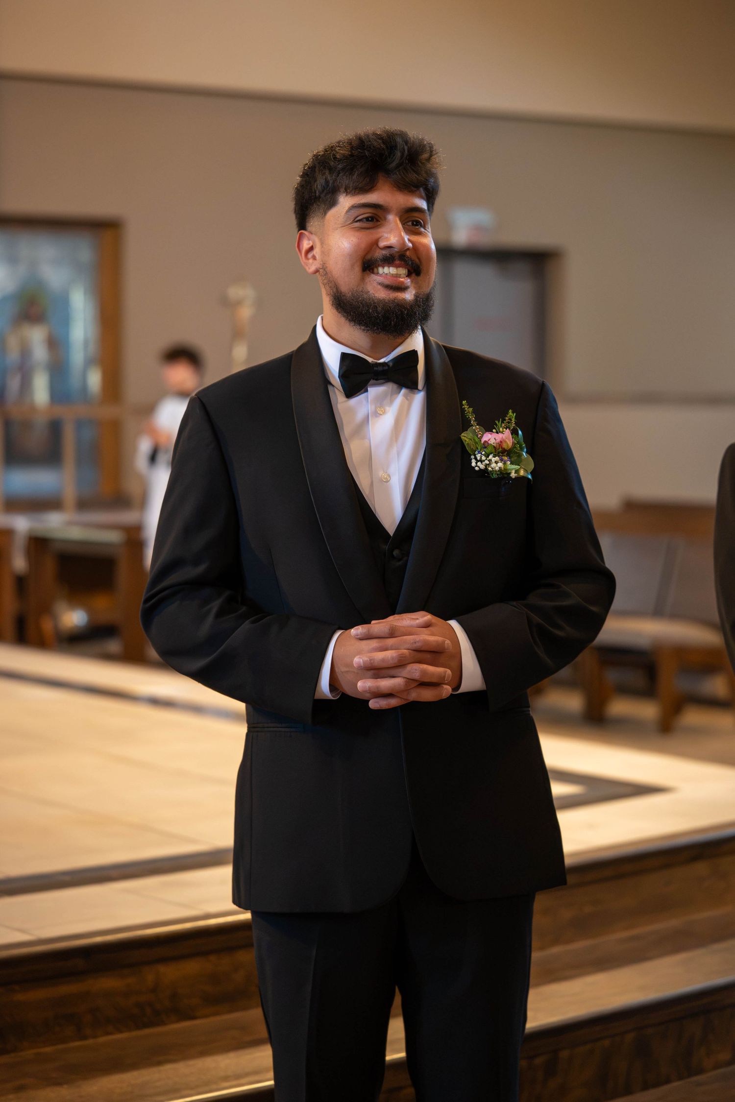Smiling groom in black tuxedo with boutonniere stands in church before wedding ceremony.