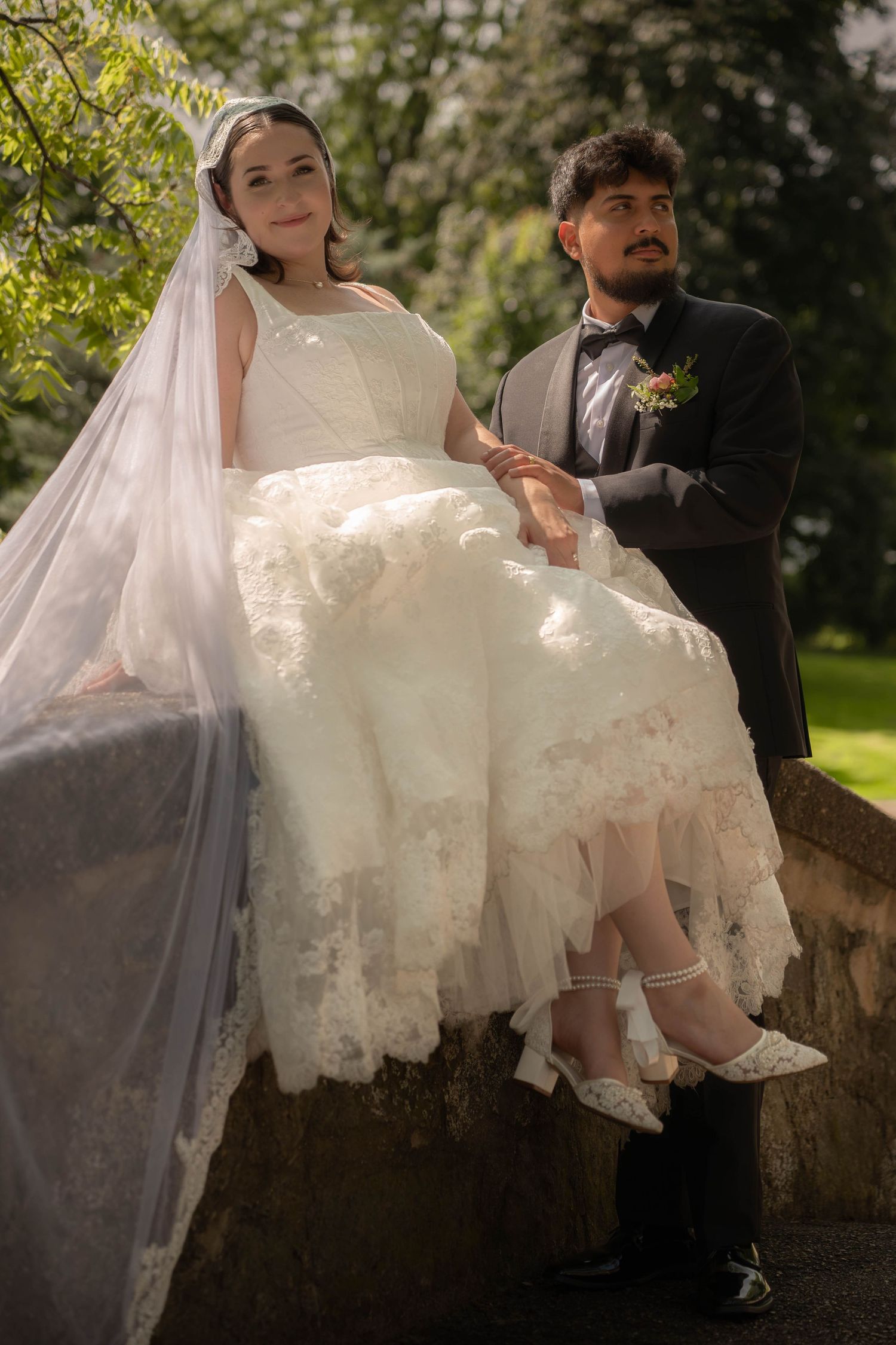 Bride in layered lace wedding dress and veil sits on stone ledge beside groom in black tuxedo in sunny park.