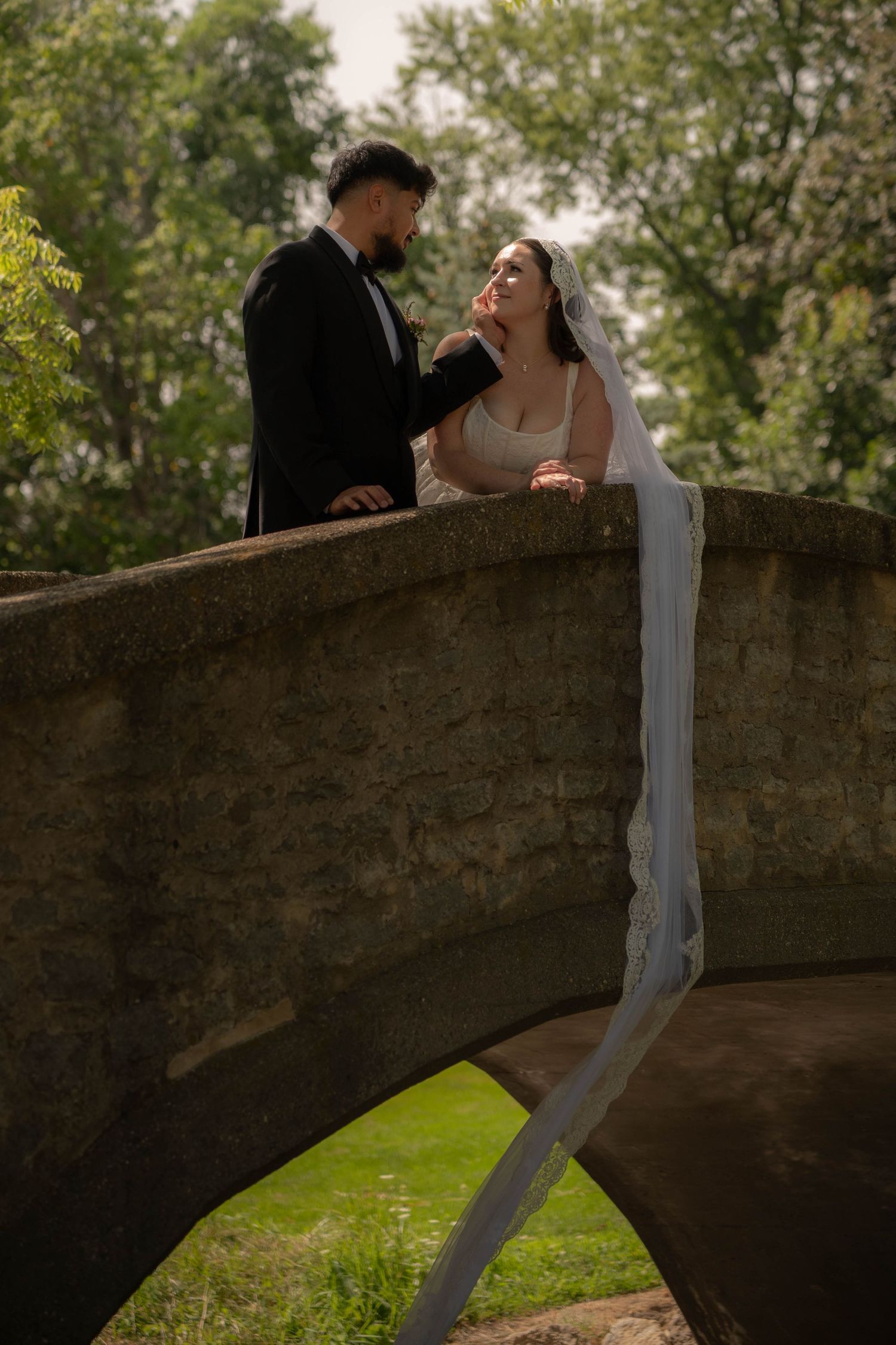 Bride in white lace gown and groom in black tuxedo pose together on a rustic stone arched bridge.