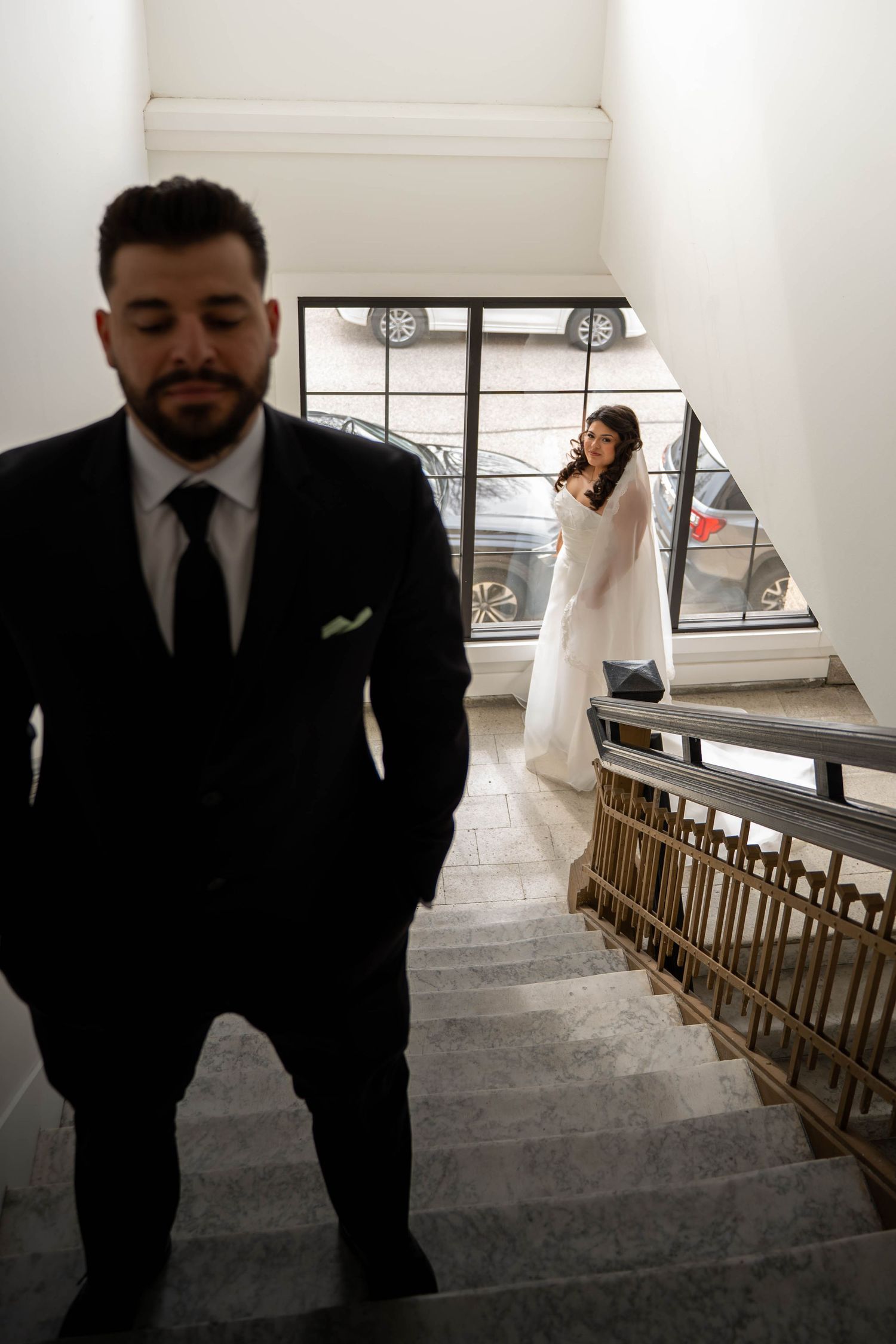 Groom in black suit descends stairs as bride in white gown watches from above.