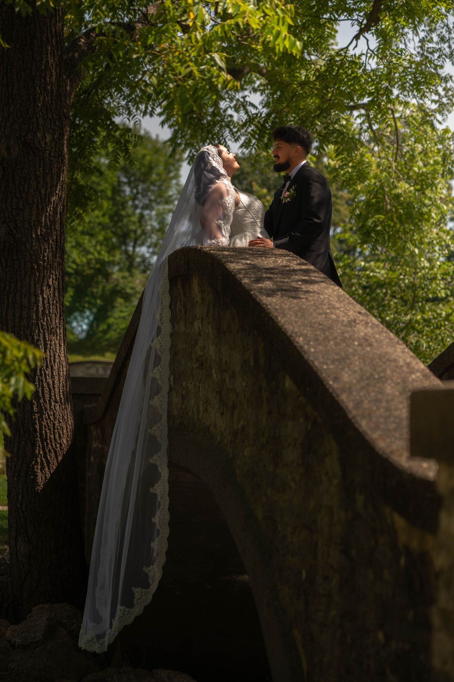Bride in lace veil and groom in black tuxedo share a romantic moment on a stone bridge outdoors.
