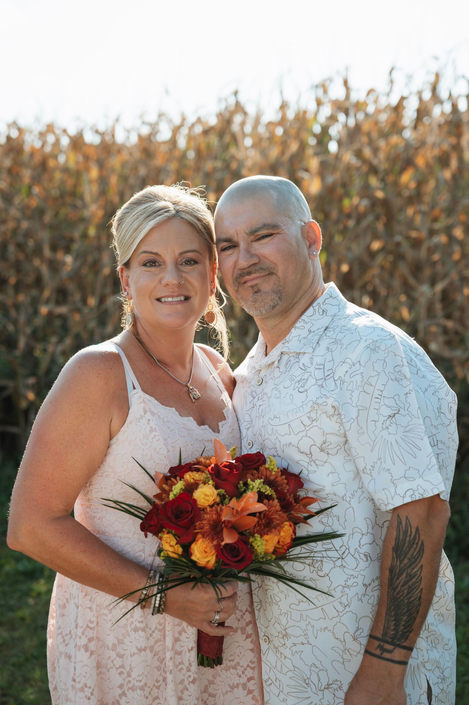 Wedding couple posing before cornfield, bride holding autumn bouquet of red and orange flowers.