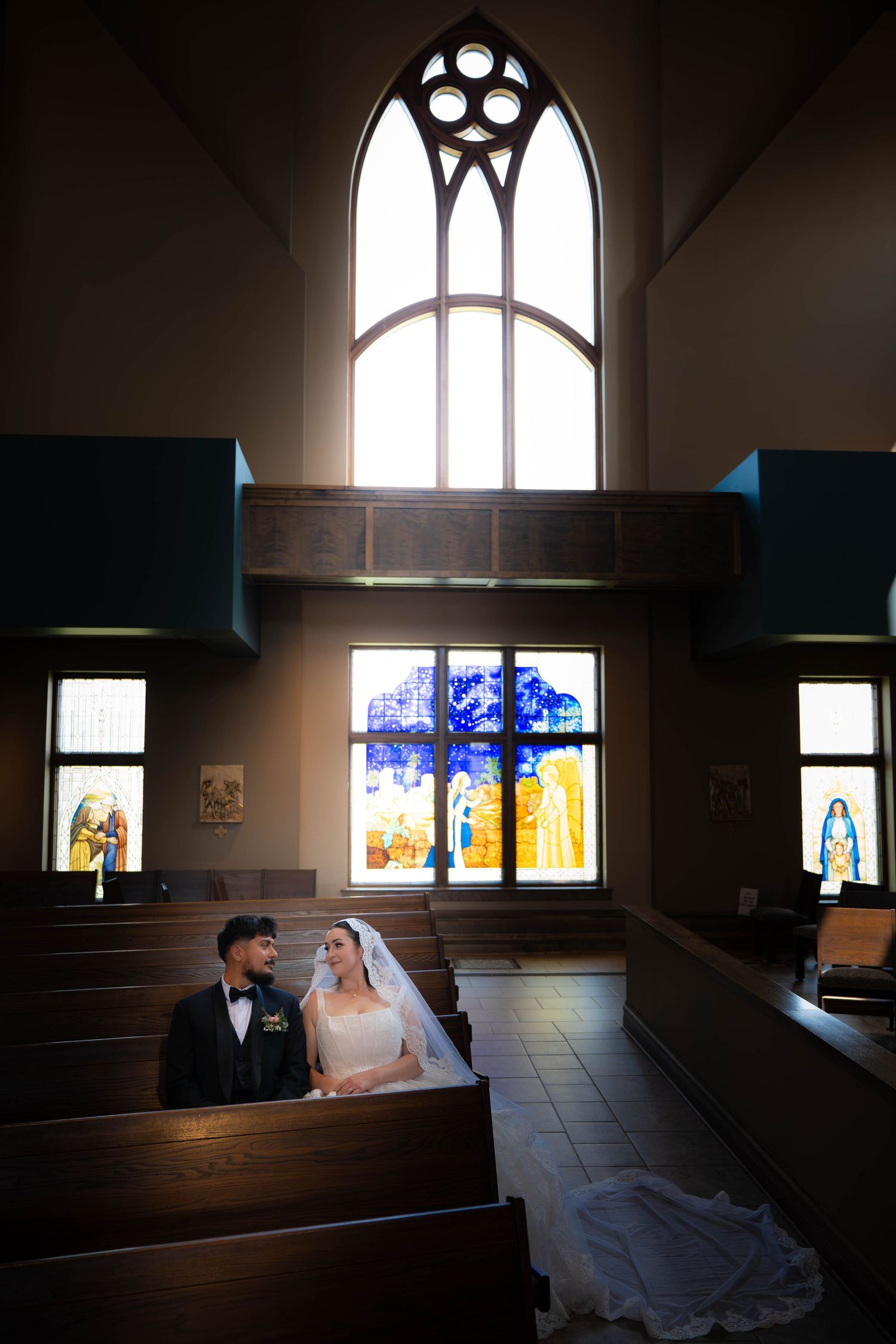 A bride and groom sitting together in a sunlit church pew with stained glass windows above.