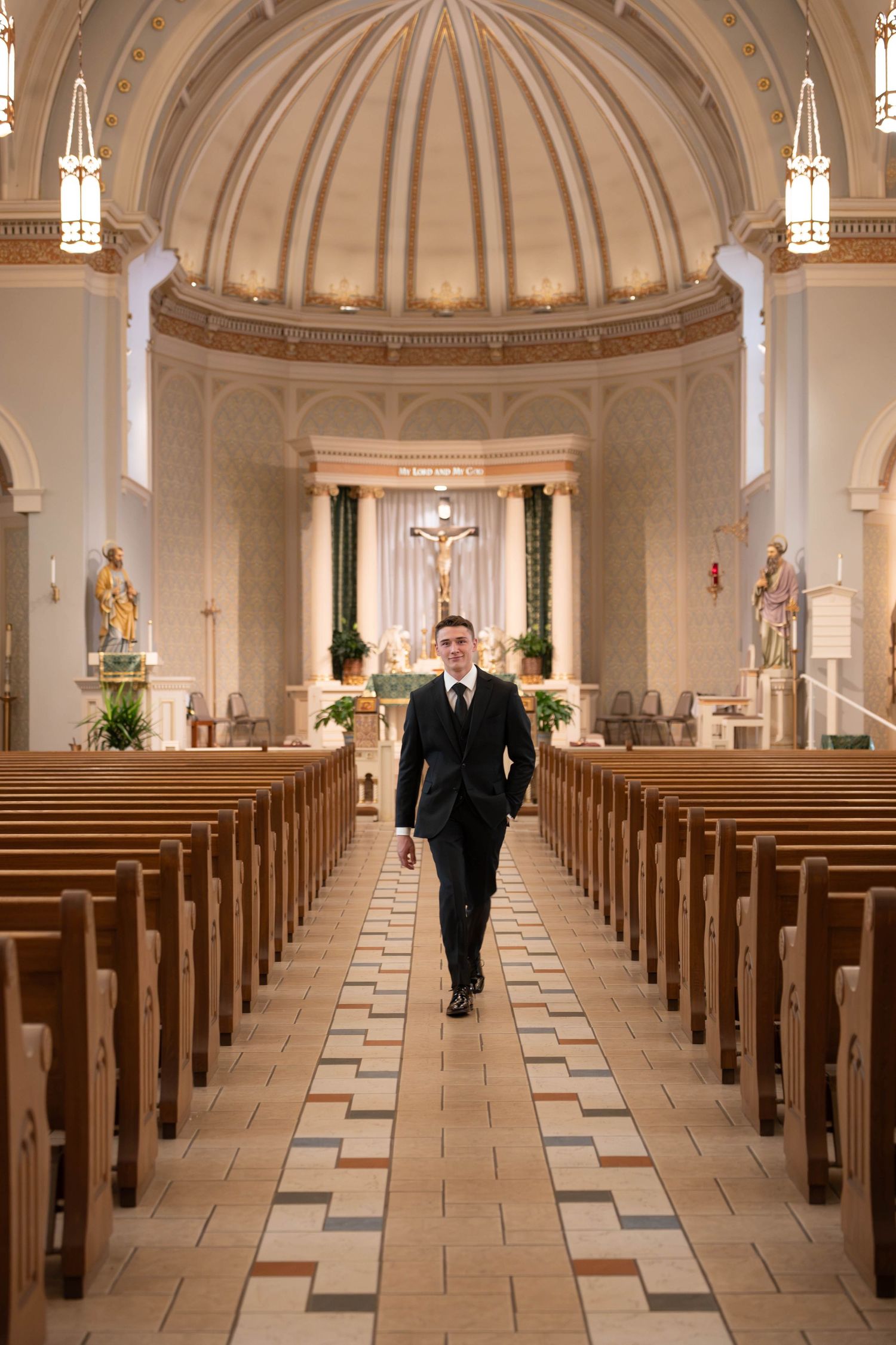 Groom in black suit walking down aisle of elegant domed Catholic church before wedding ceremony.